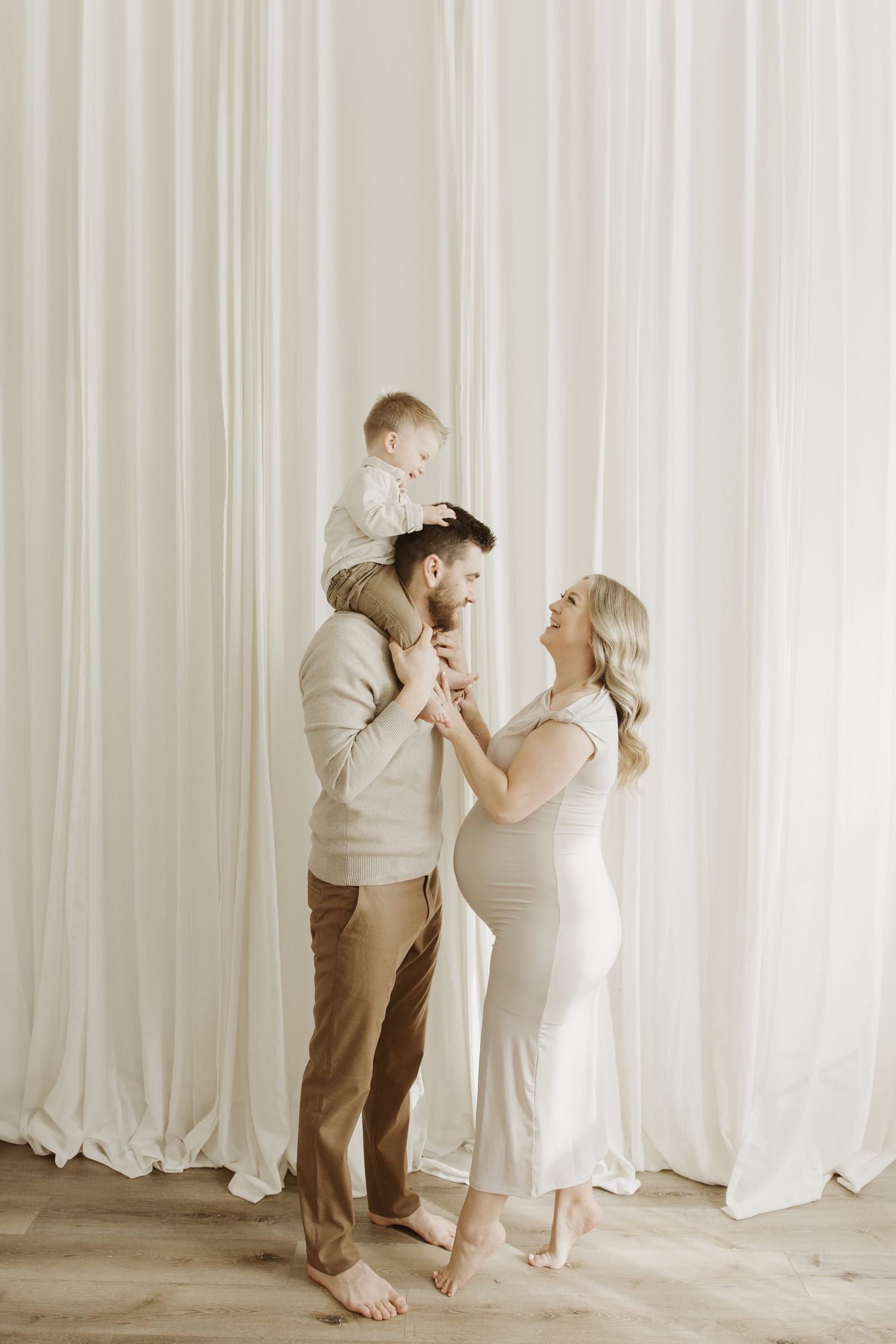 Happy family with pregnant mother, father, and baby on shoulders posing in front of white curtains.