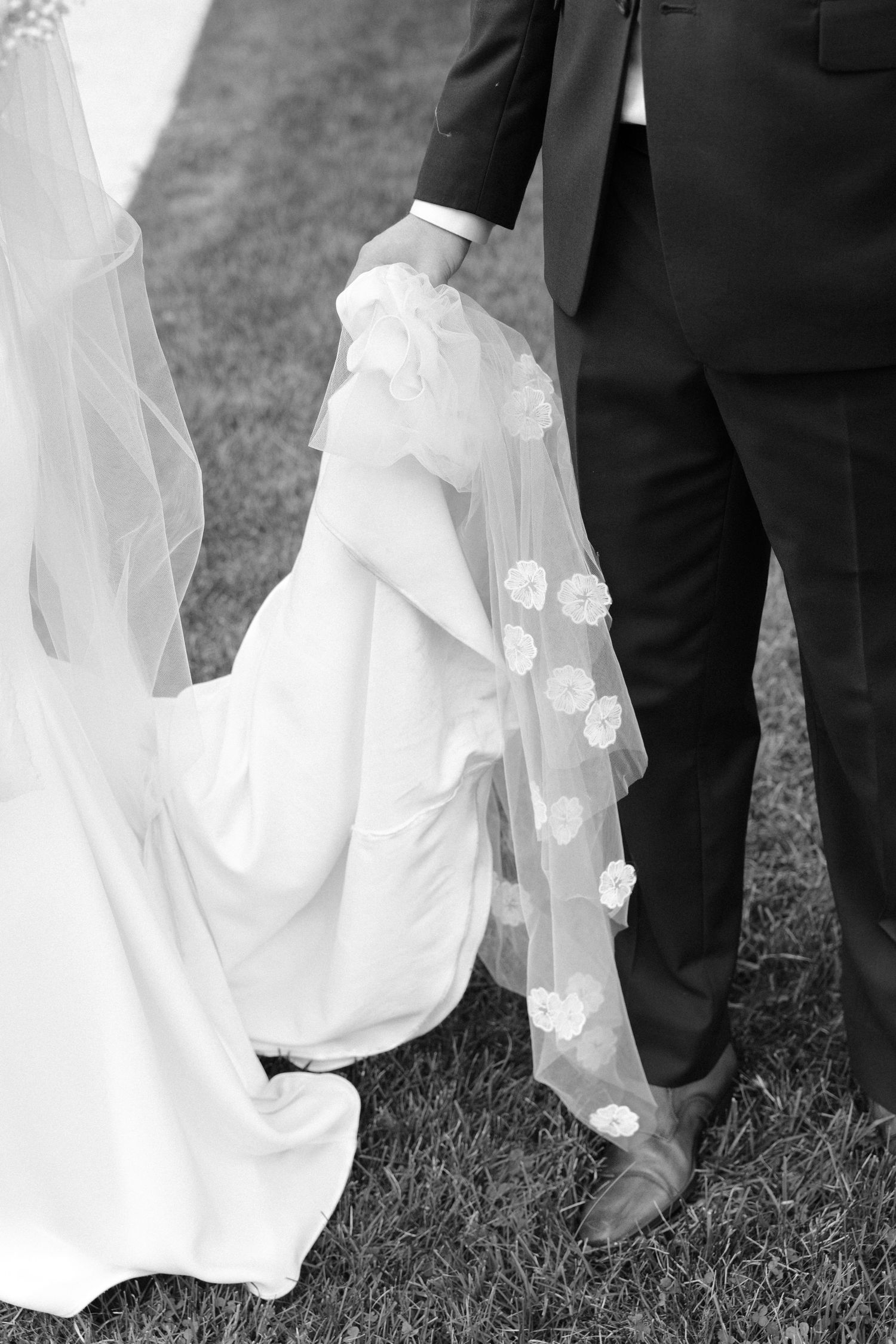 Black and white close-up of bride and groom holding hands, showcasing the bride's floral-trimmed wedding veil.