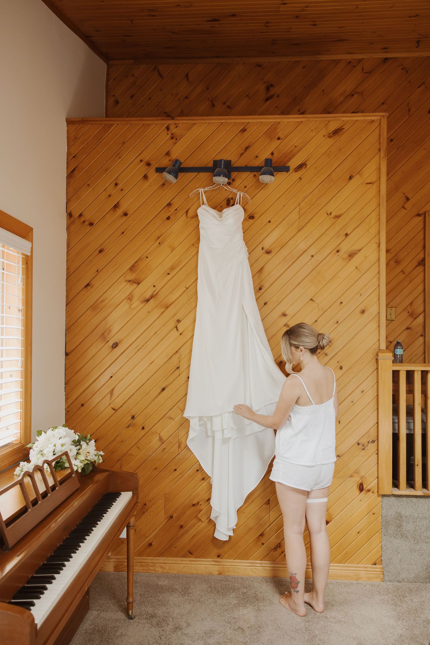 Bride in white camisole admires her wedding dress hanging on wooden wall above piano.