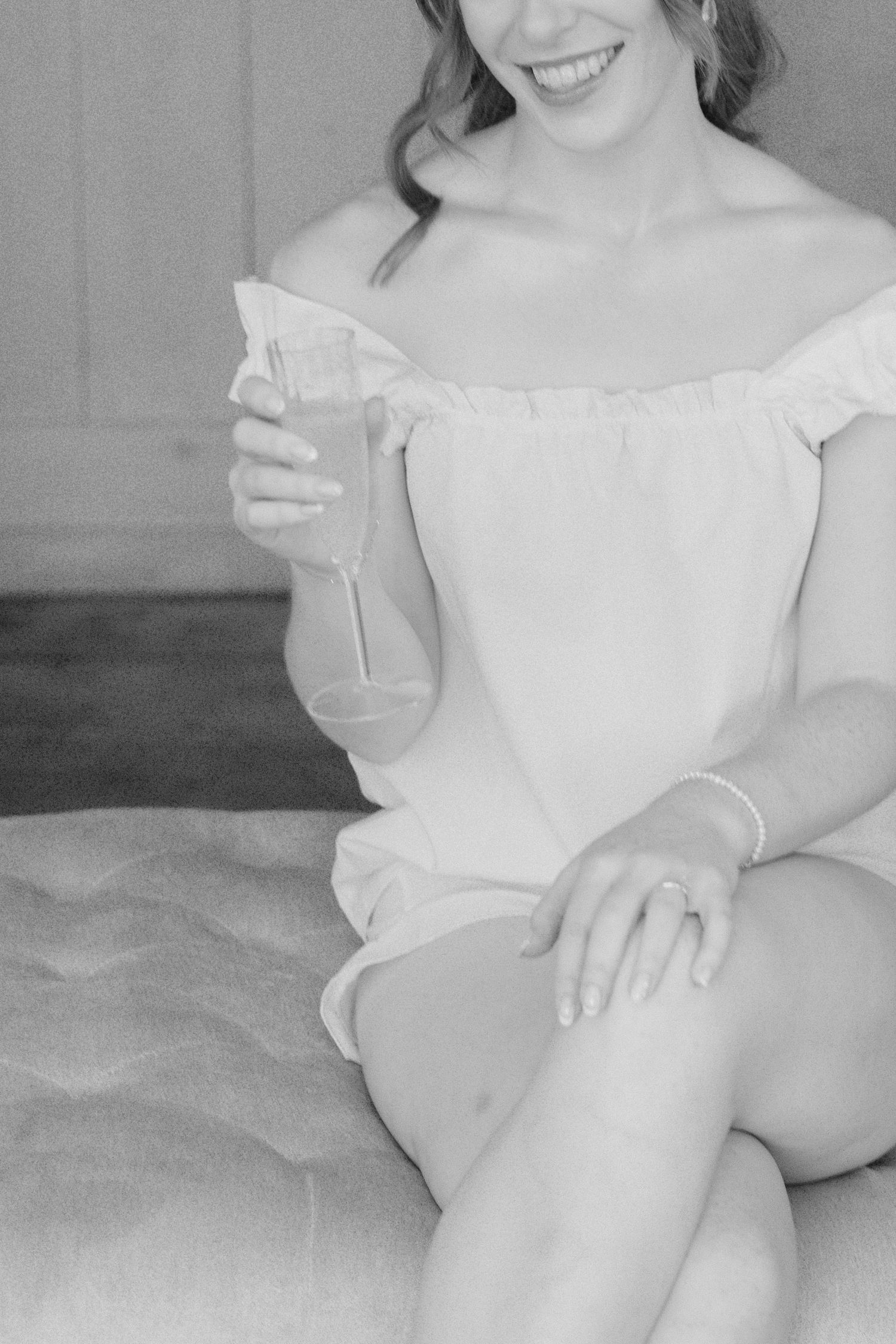 Black and white close-up of a smiling bride in an off-shoulder top holding a champagne glass while seated.