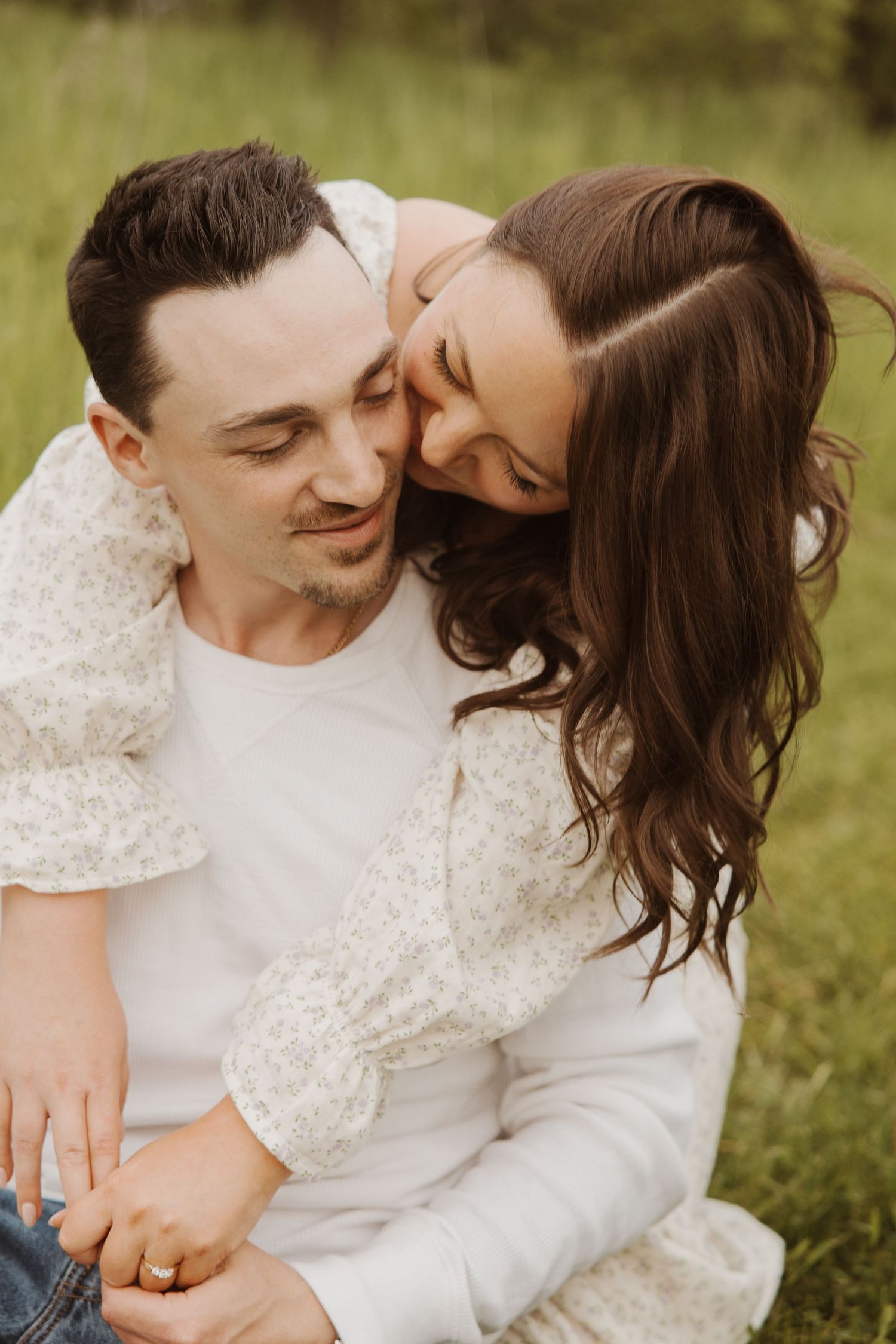 Woman in floral dress resting her head on man's cheek while sitting together in a lush green field.