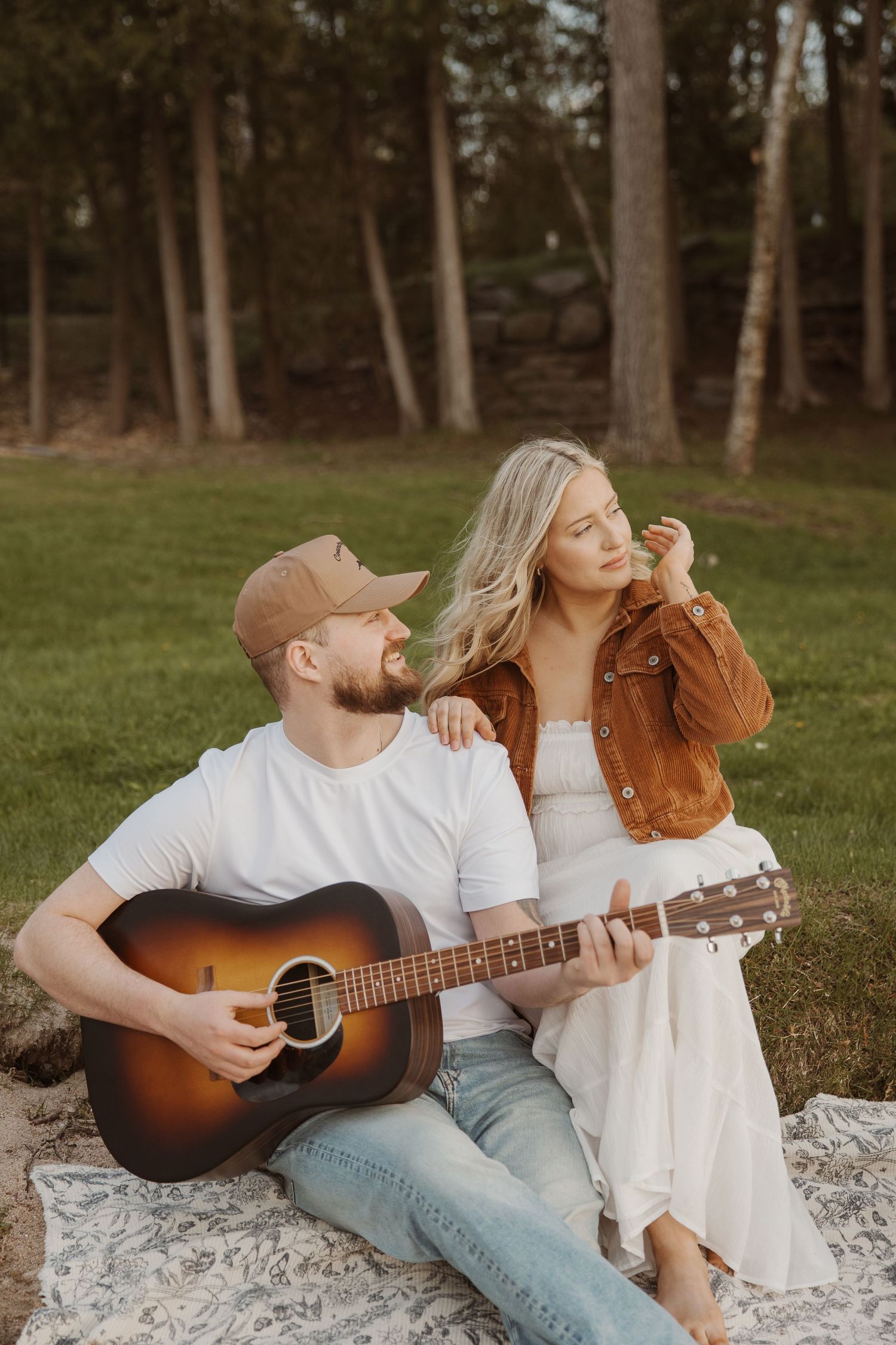 Man playing acoustic guitar while woman in white dress leans on him outdoors.