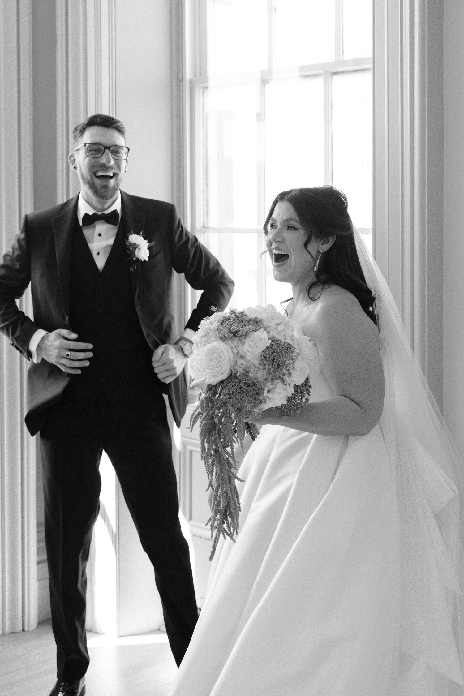 Black and white photo of joyful bride and groom laughing together by a bright window after ceremony.