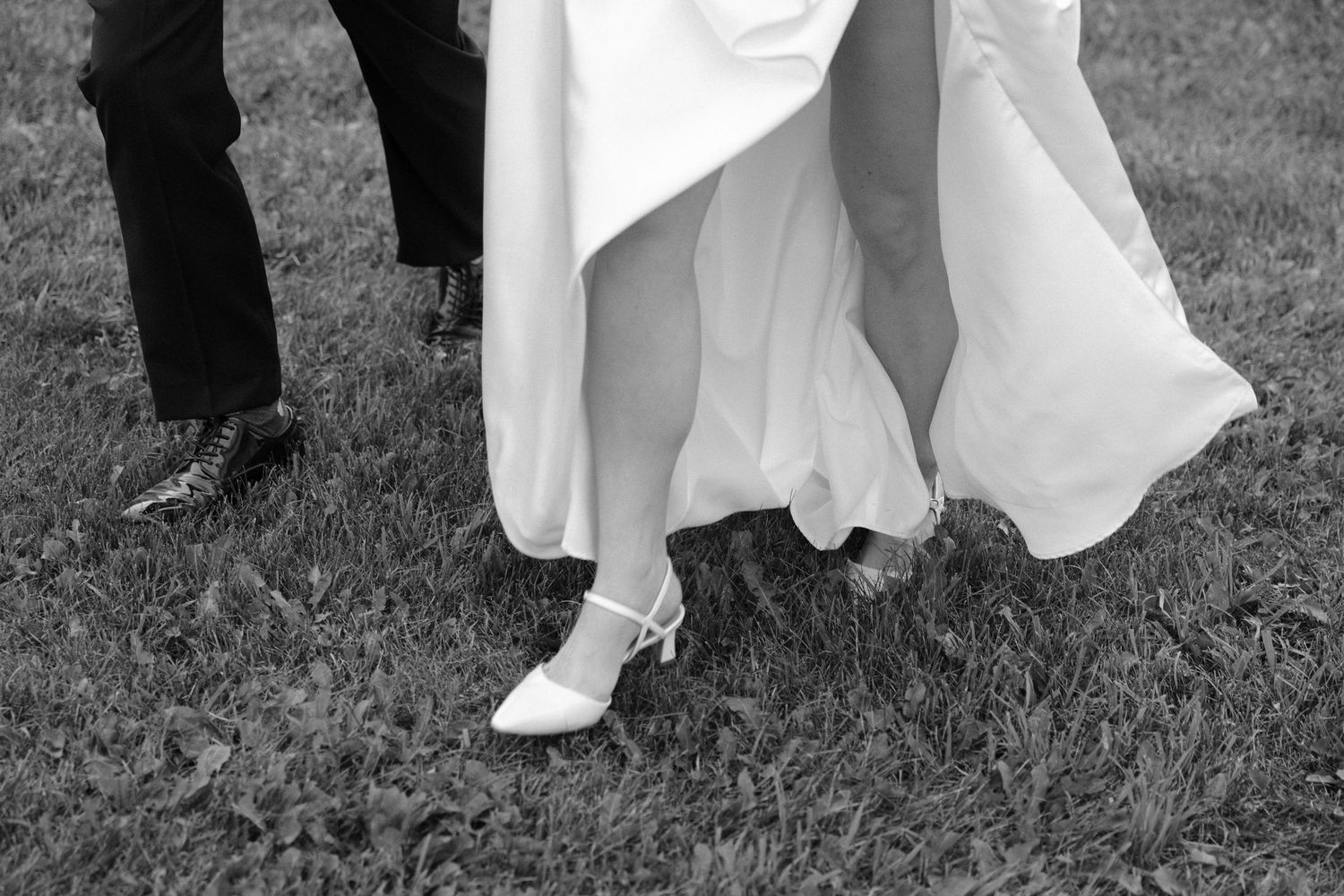 Black and white photo of bride in white gown and heels walking on grass with groom.