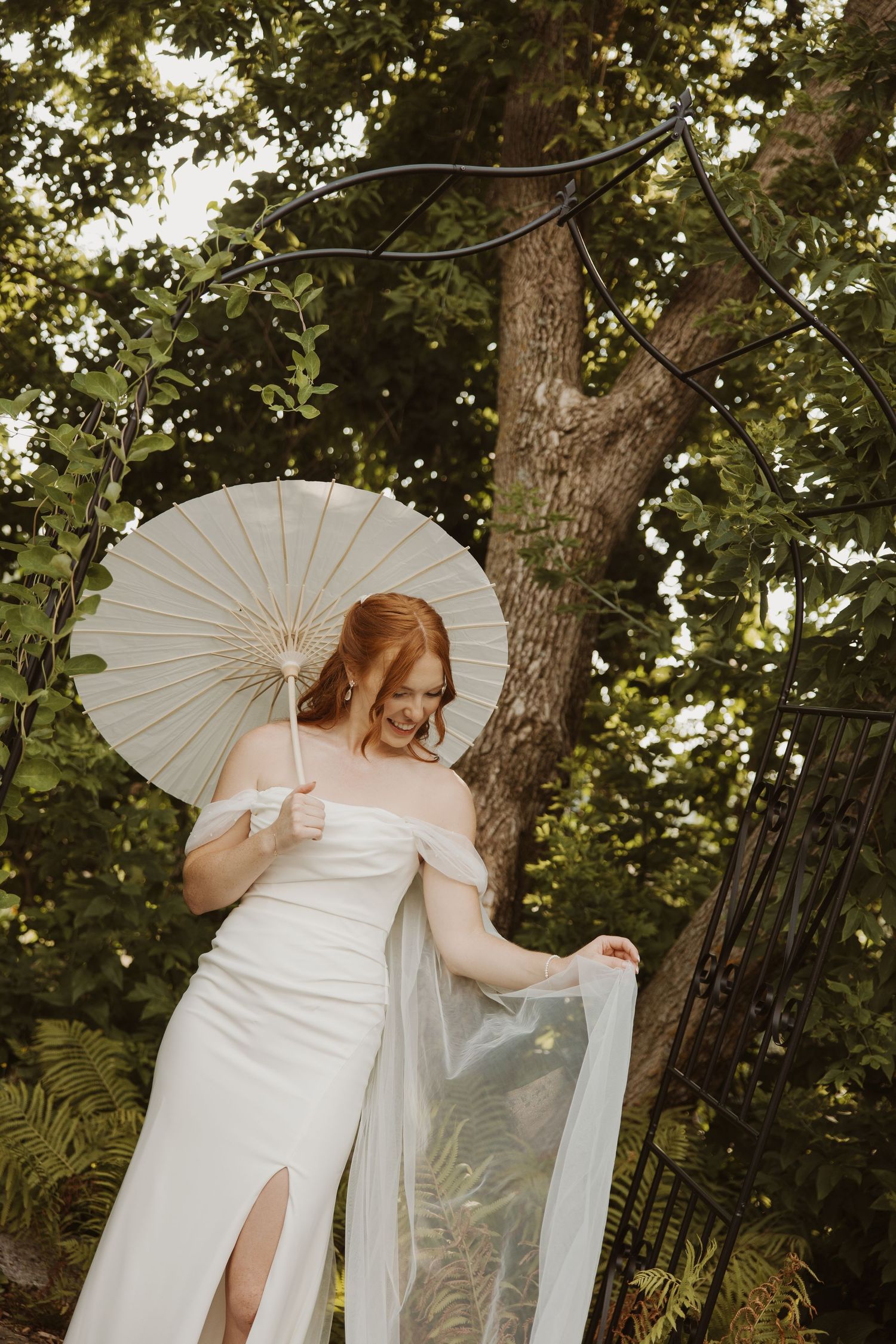 A radiant bride in a white off-shoulder gown holds a parasol while posing in a lush garden.