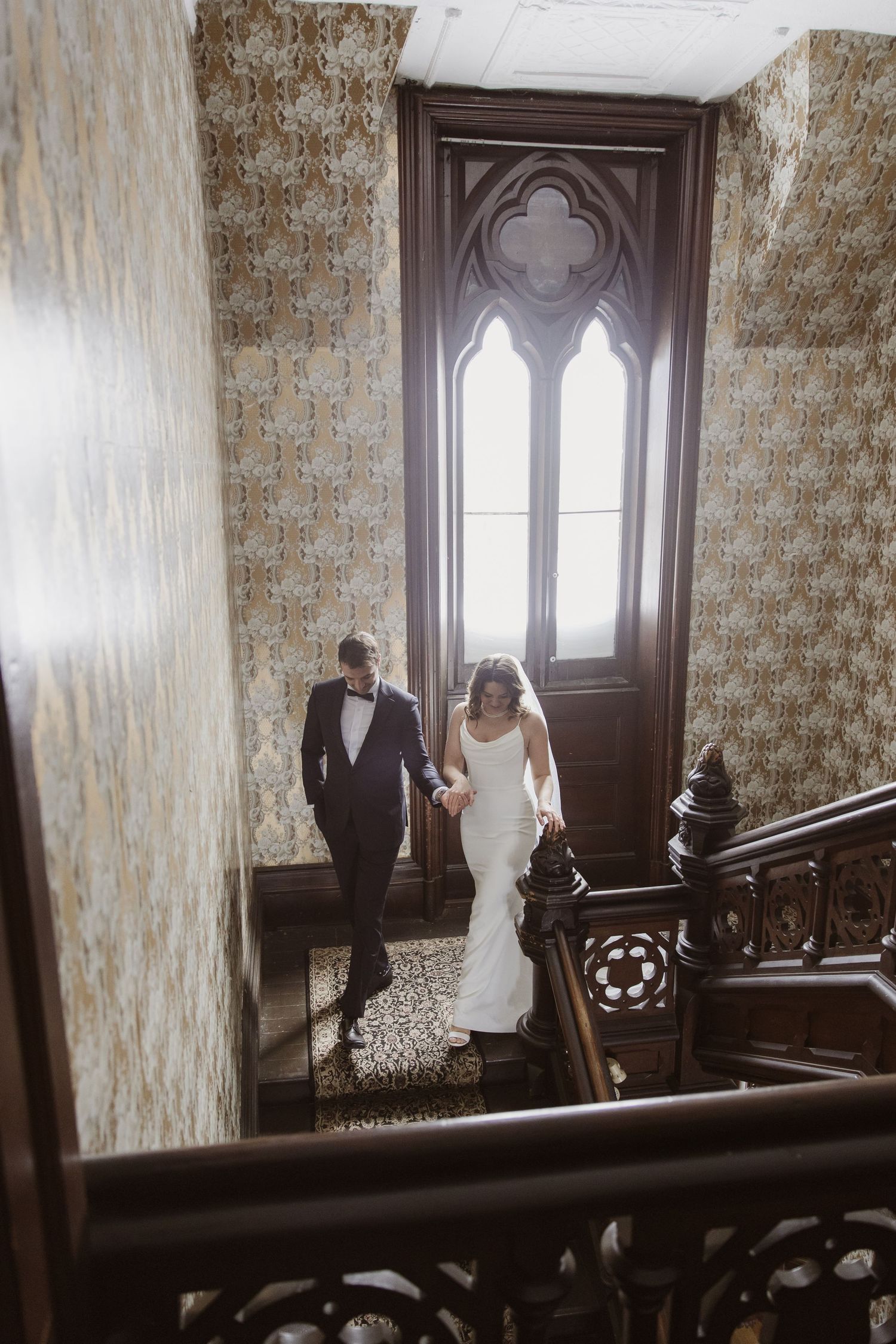 Bride and groom holding hands while descending an ornate wooden staircase in a historic wedding venue.