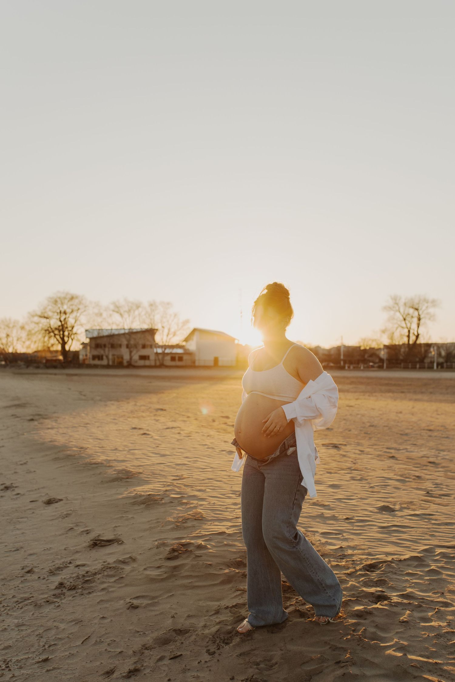 Pregnant woman cradling her baby bump on a sandy beach at golden hour sunset.