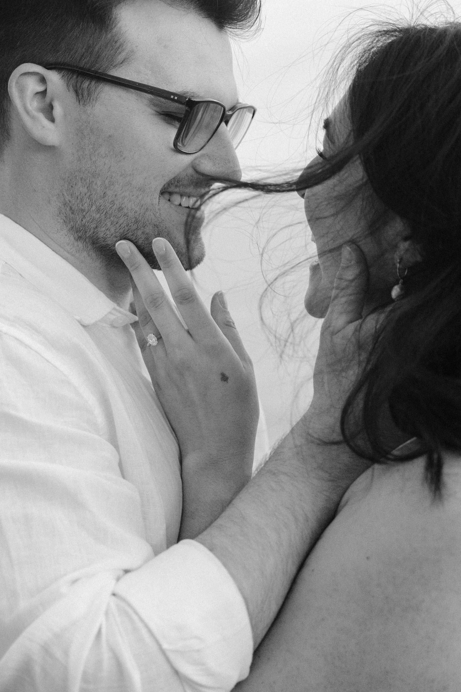 Black and white close-up of smiling couple almost kissing, engagement ring visible.