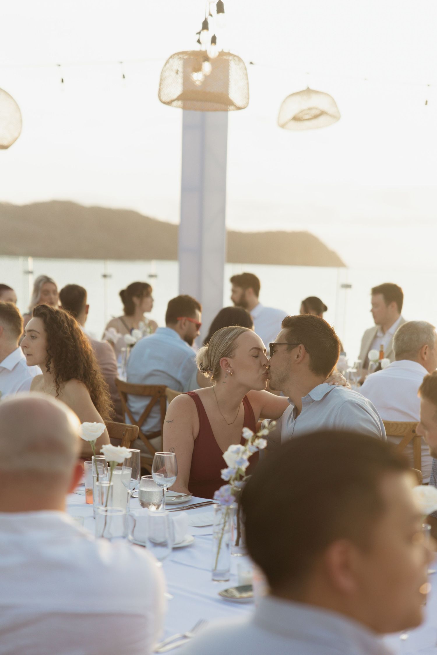 Couple sharing a kiss at an outdoor beachside wedding reception dinner at sunset with guests seated around them.