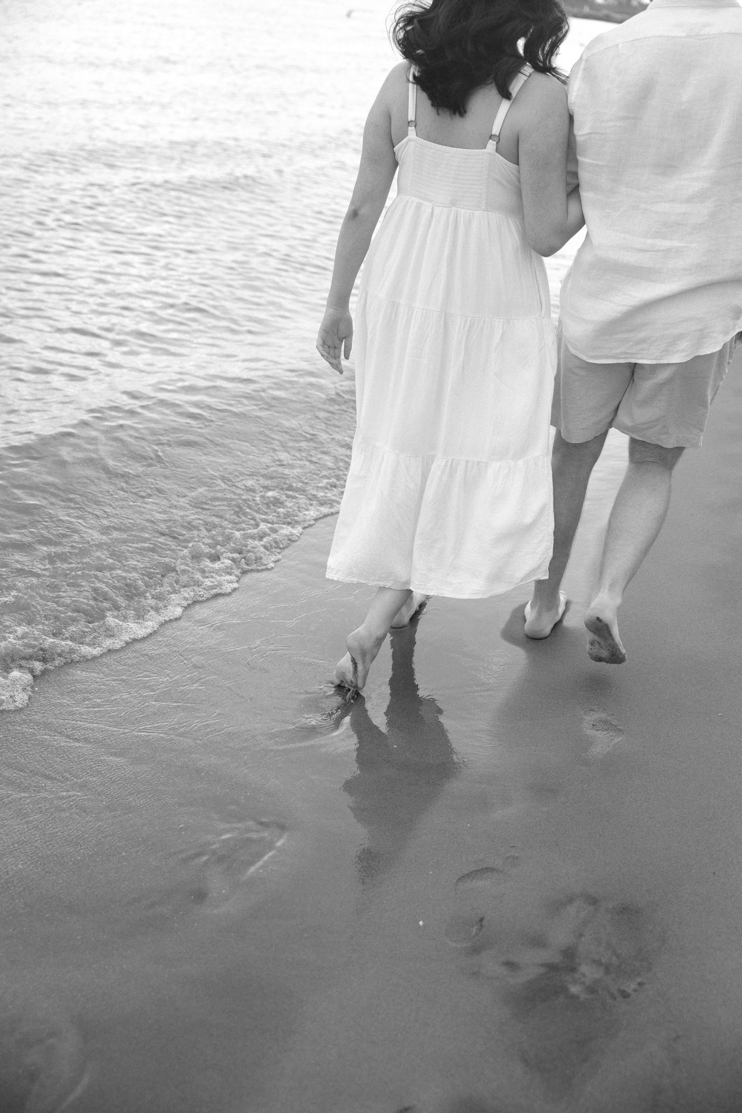Black and white photo of couple walking barefoot along ocean shoreline together.