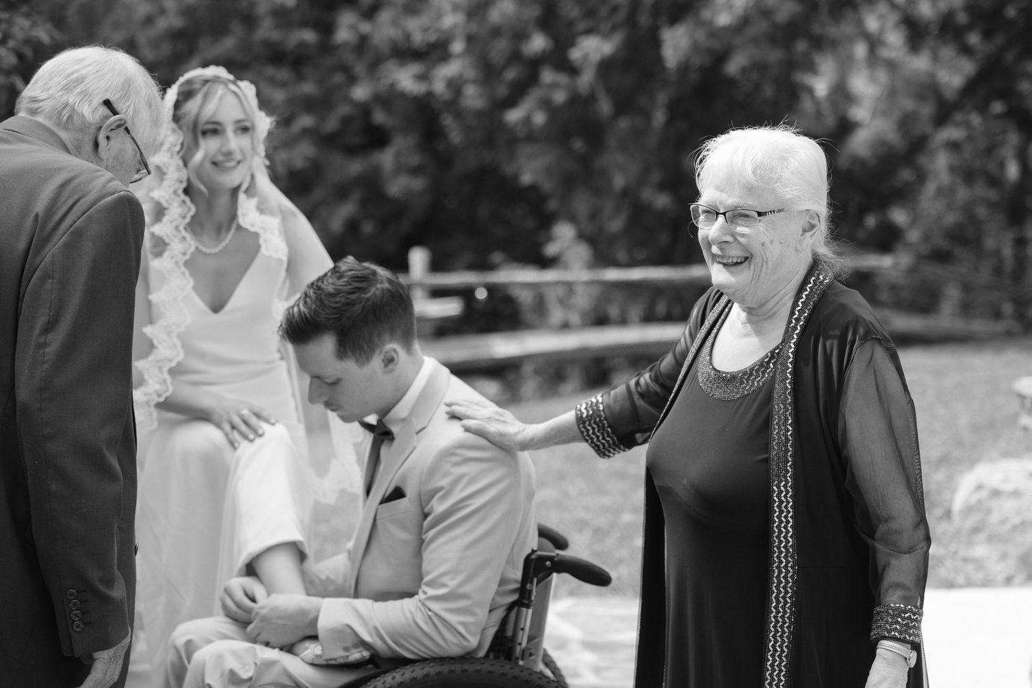 Groom in wheelchair greeted by elderly woman during outdoor wedding reception.