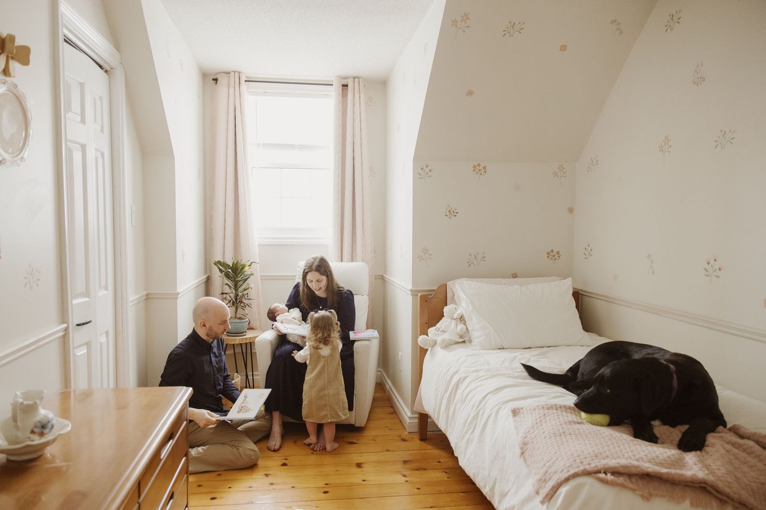 Family spending quality time in a cozy bedroom with floral wallpaper, a child, parents, and a black dog.