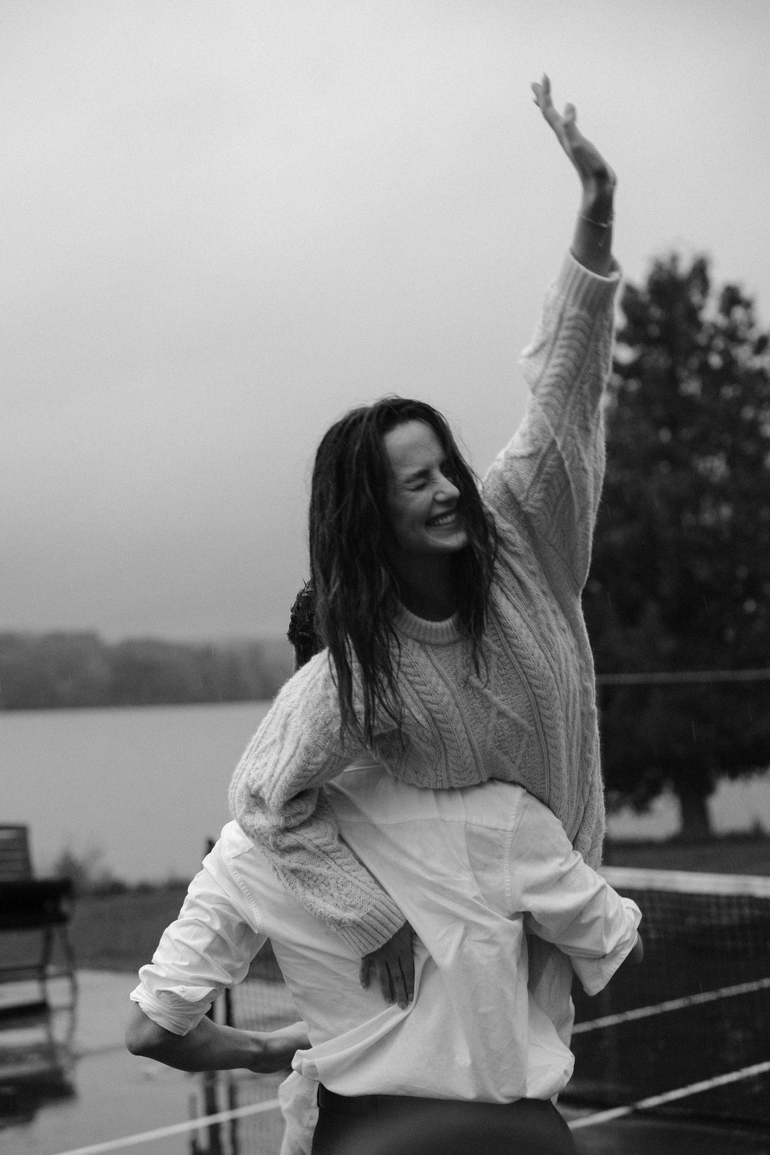 Black and white photo of joyful woman raising arm on rainy lakeside deck.