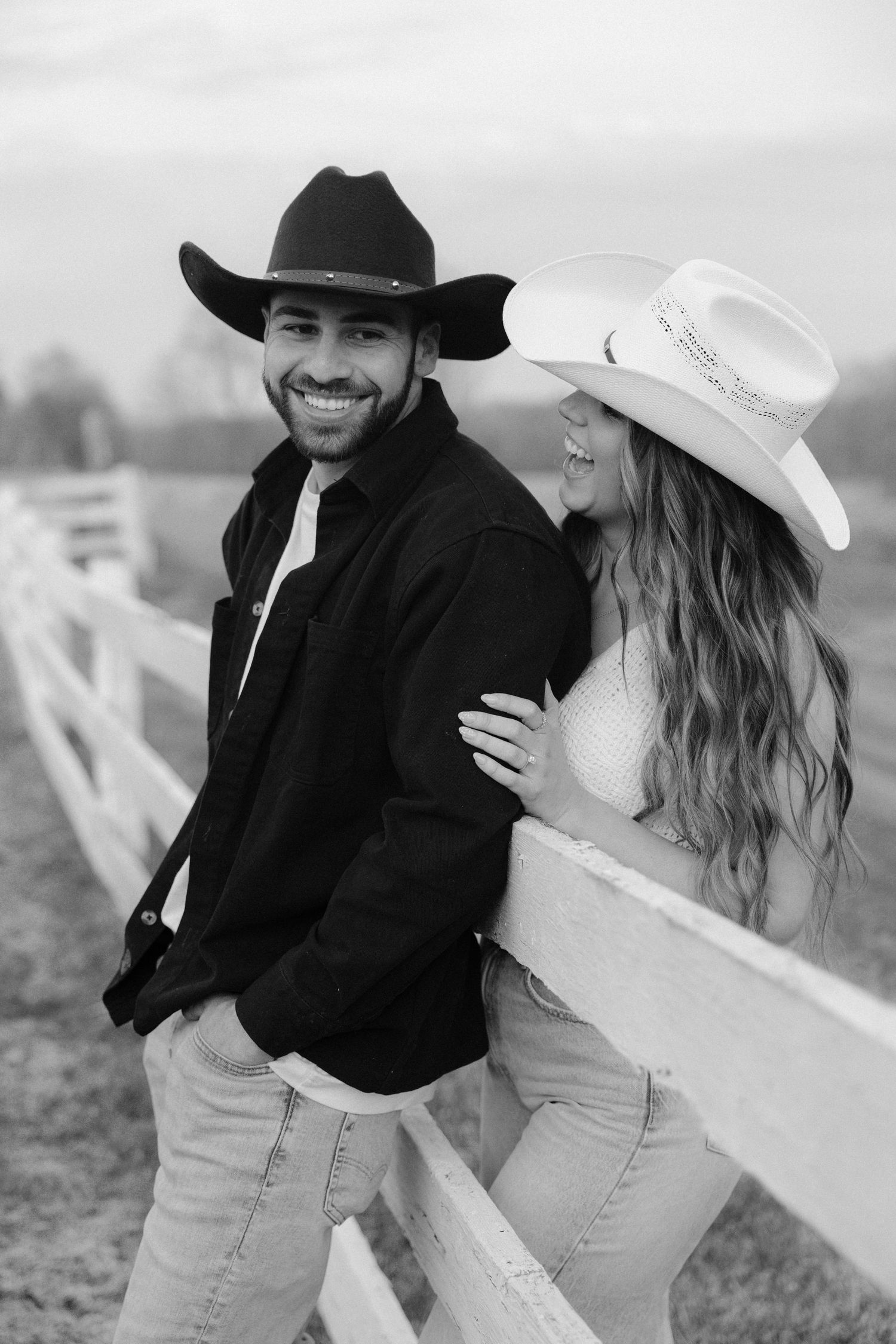 Smiling couple in cowboy hats leaning against white fence on ranch in black and white.