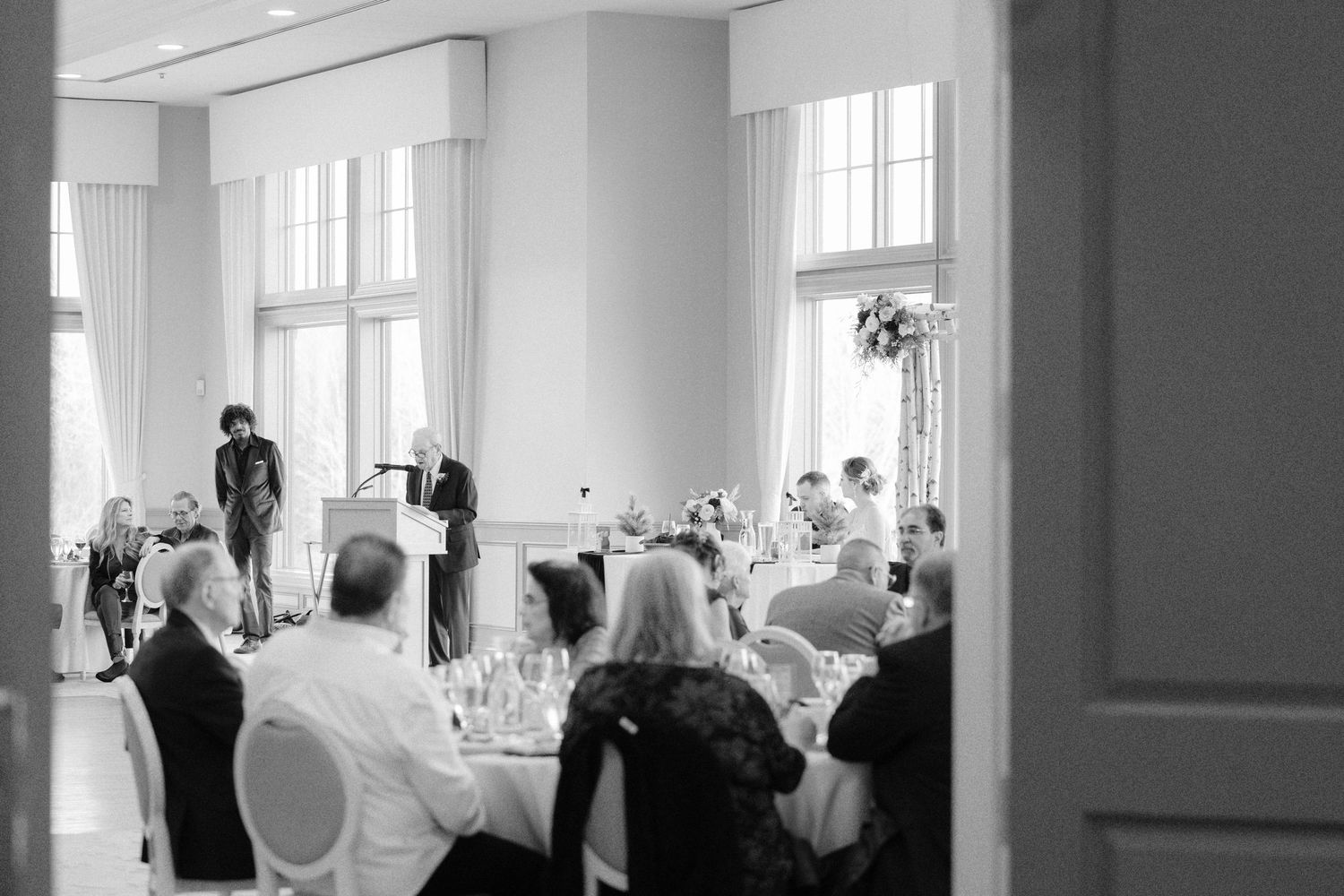 Black and white wide shot of wedding reception guests seated at tables in an elegant ballroom venue.