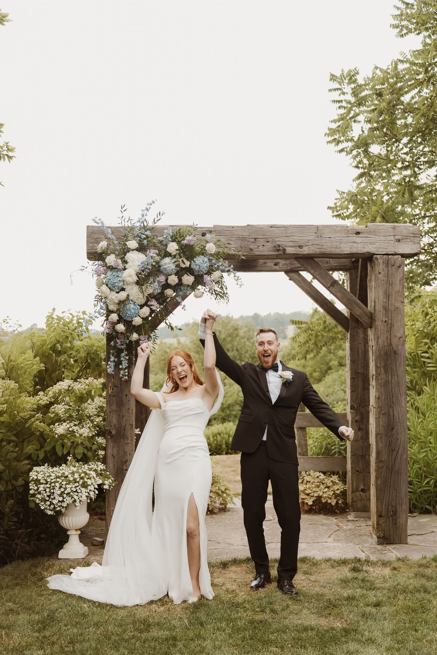 Joyful newlywed couple raising hands under rustic wooden arch adorned with blue and white flowers at outdoor wedding ceremony.