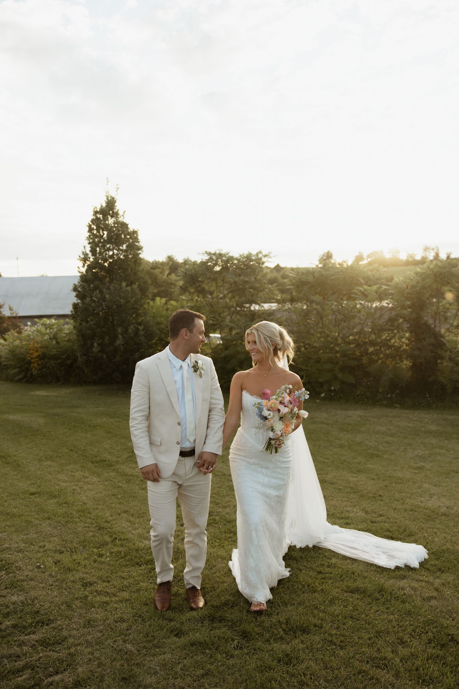 Bride and groom walk hand-in-hand on green lawn at sunset, she in white gown with bouquet, he in tan suit.