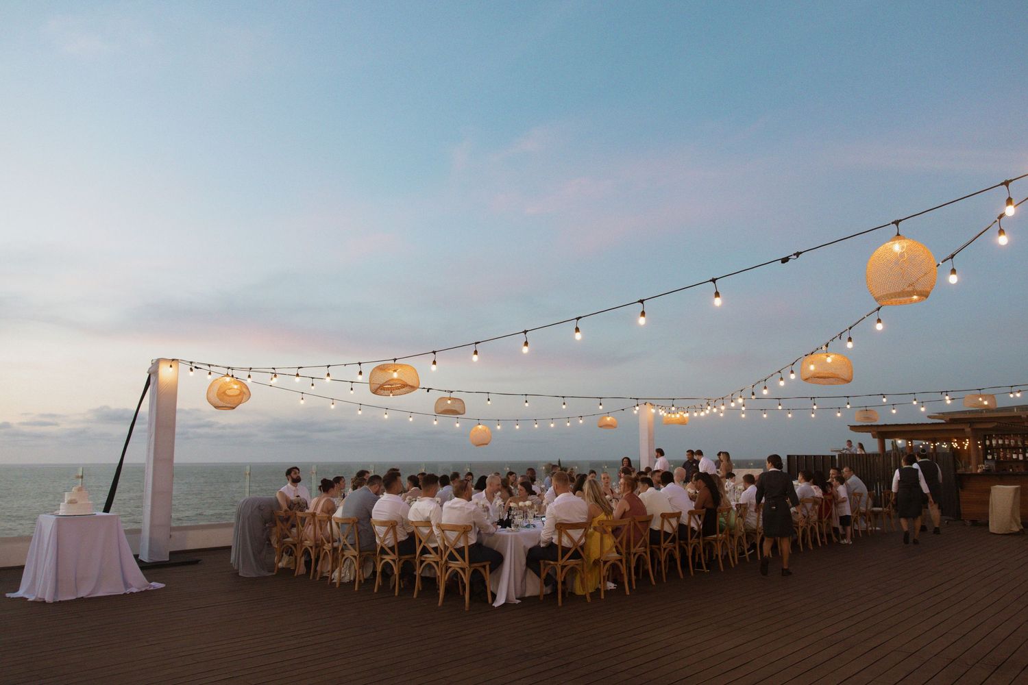 Outdoor wedding reception on oceanfront deck at dusk with string lights and paper lantern decor.