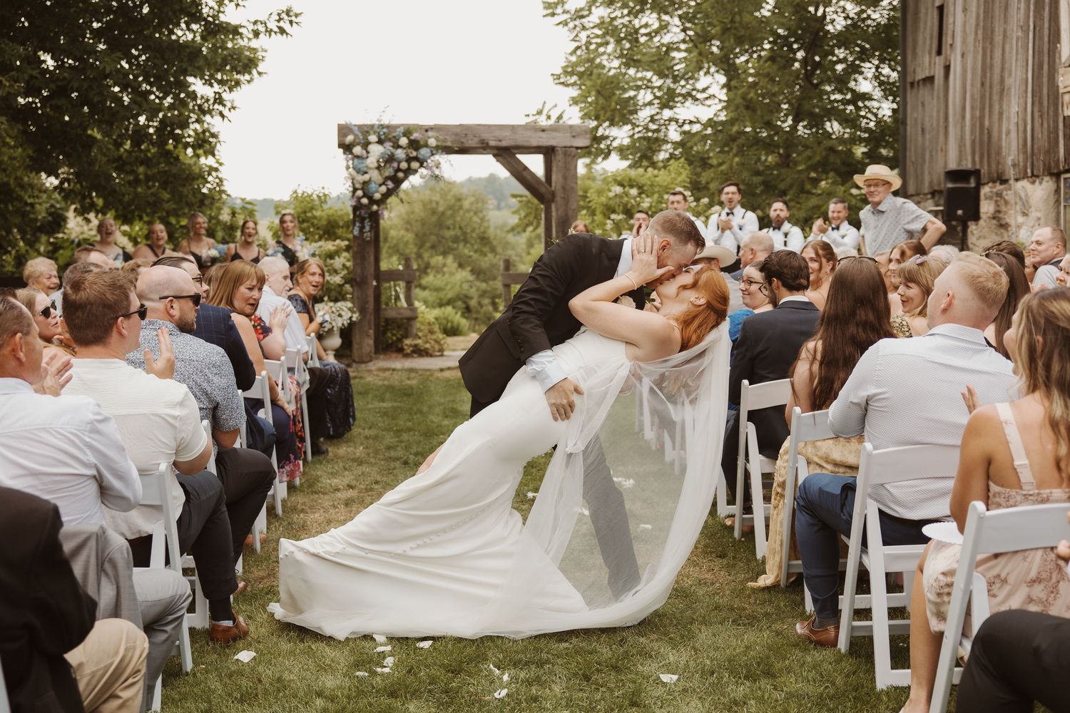 Groom dips bride for romantic kiss at outdoor wedding ceremony with seated guests watching.