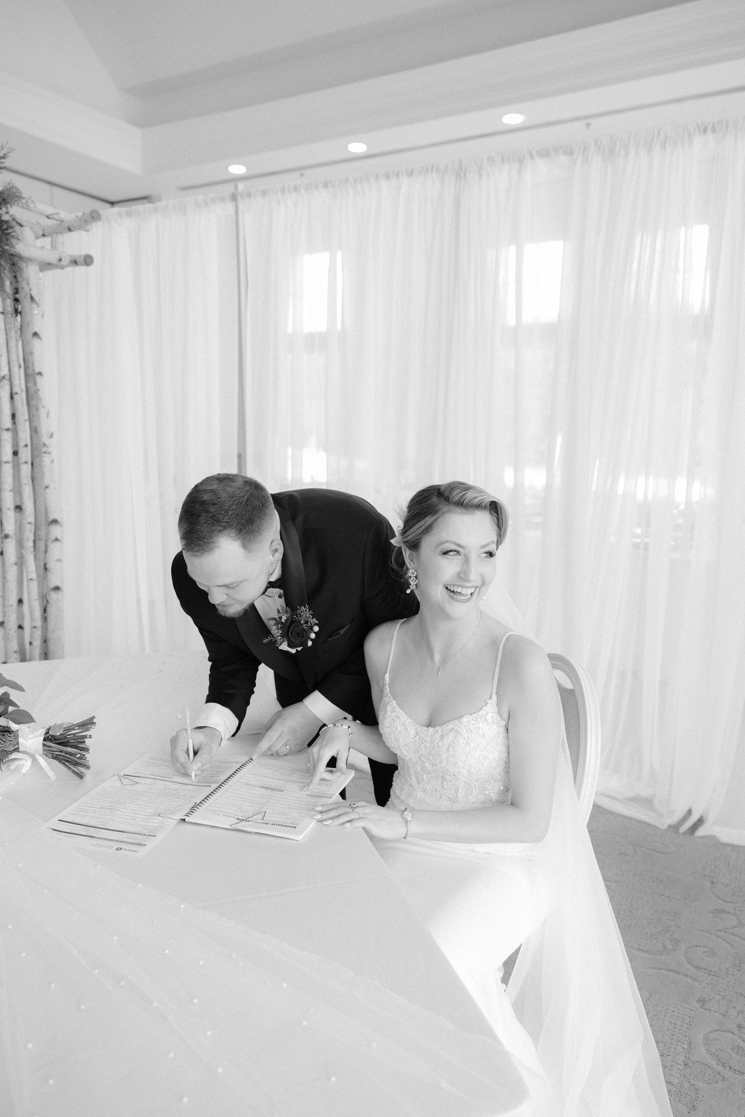 Smiling bride in white gown signing marriage certificate with groom at a table by bright windows.