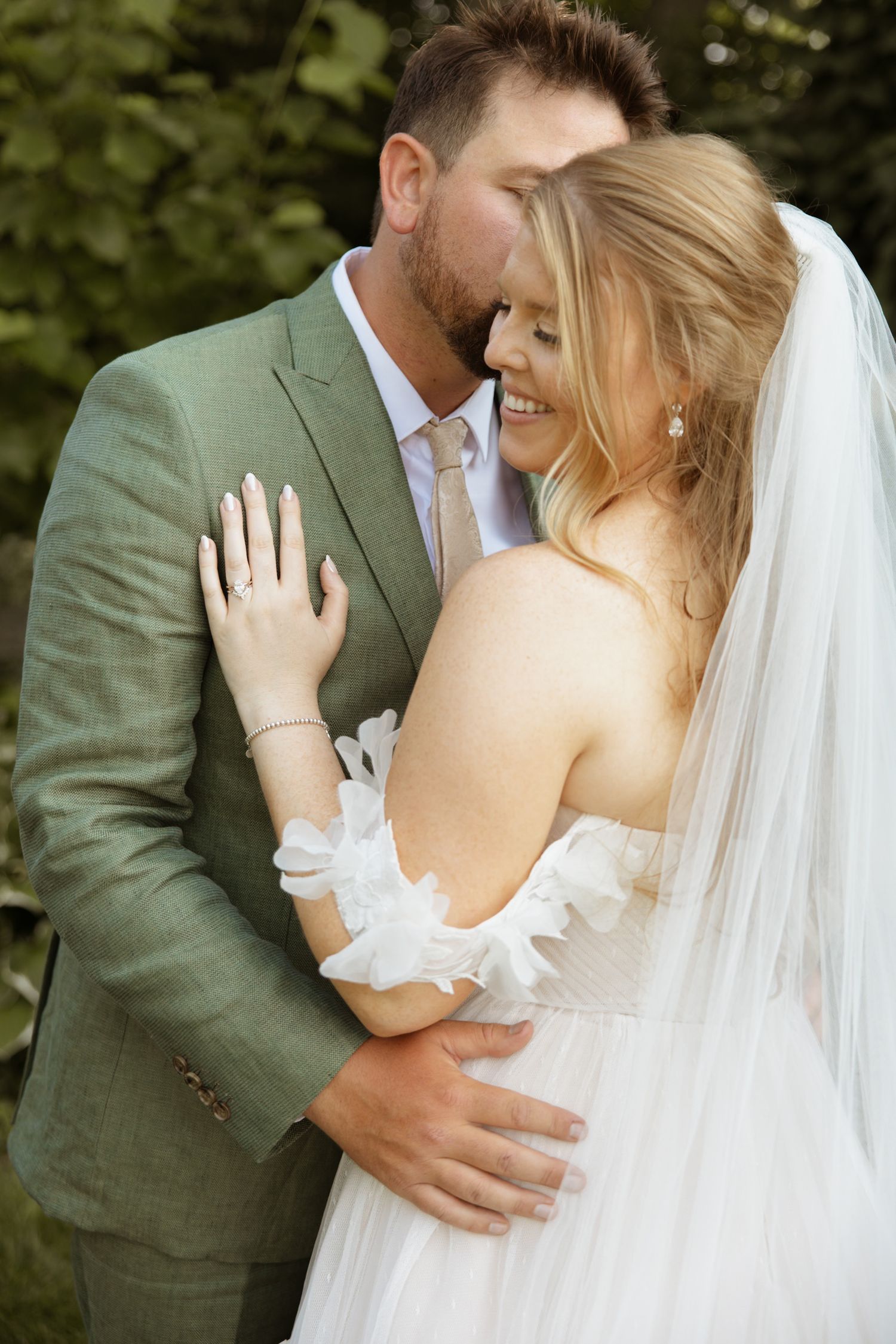 Groom in green suit embracing smiling bride in backless wedding dress and veil outdoors.