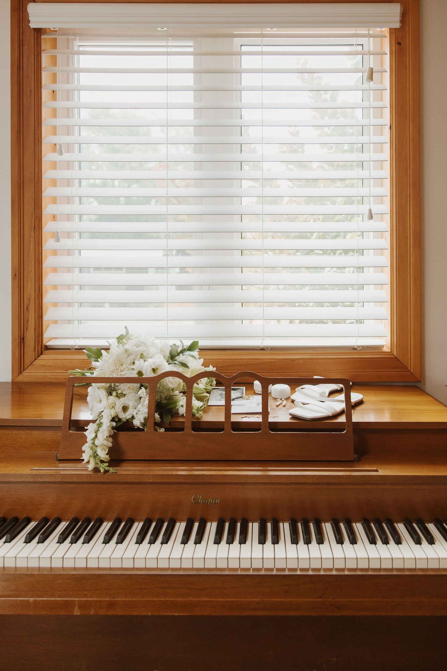 White wedding bouquet and rings resting on wooden piano keys beside a sunlit window.
