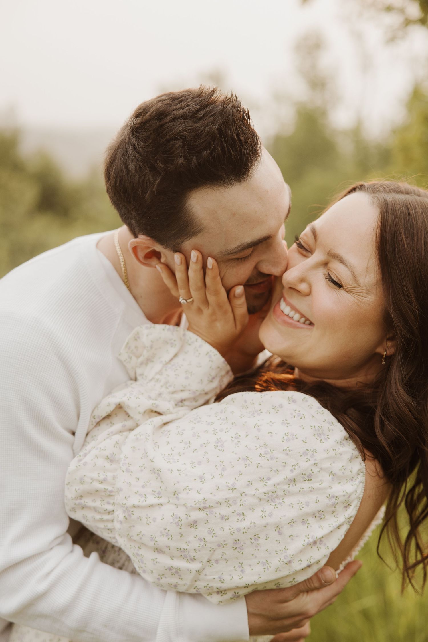Man tenderly kissing smiling woman's cheek while embracing her outdoors in a green field.