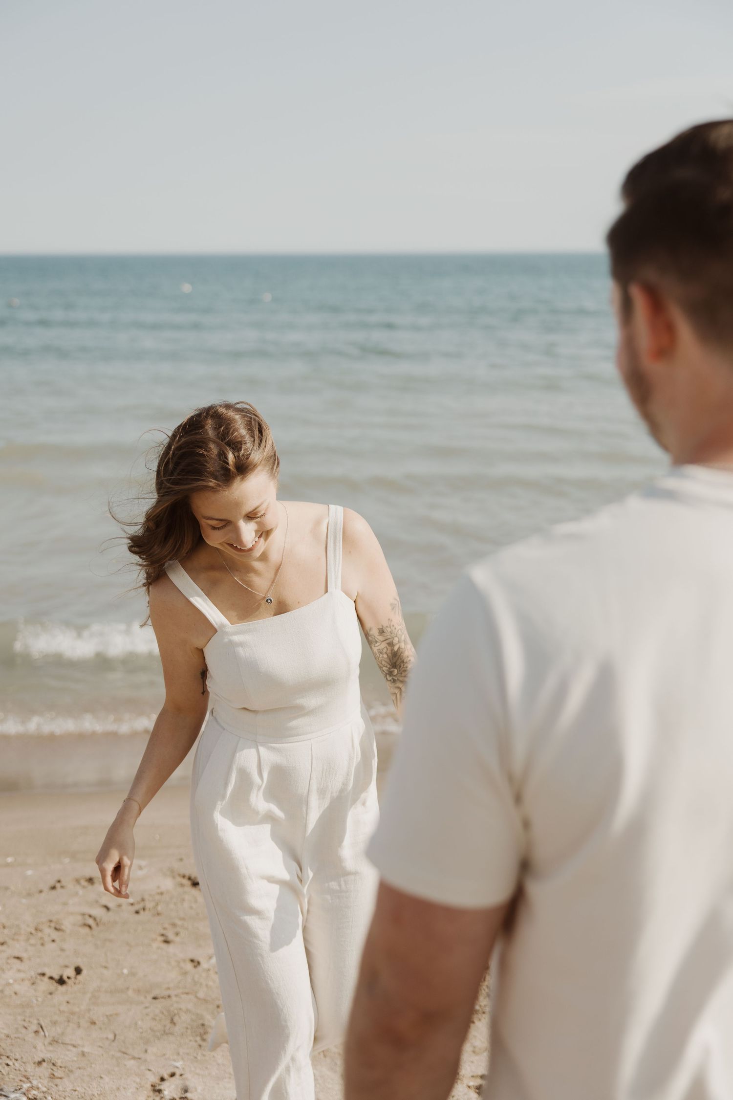Woman in white jumpsuit walking hand in hand with man along sunny ocean beach.