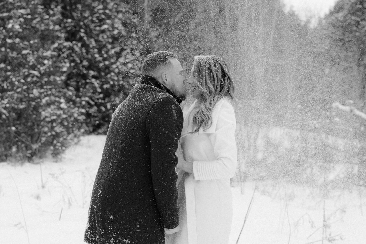 Black and white photo of a couple sharing a tender kiss in a snowy winter forest setting.