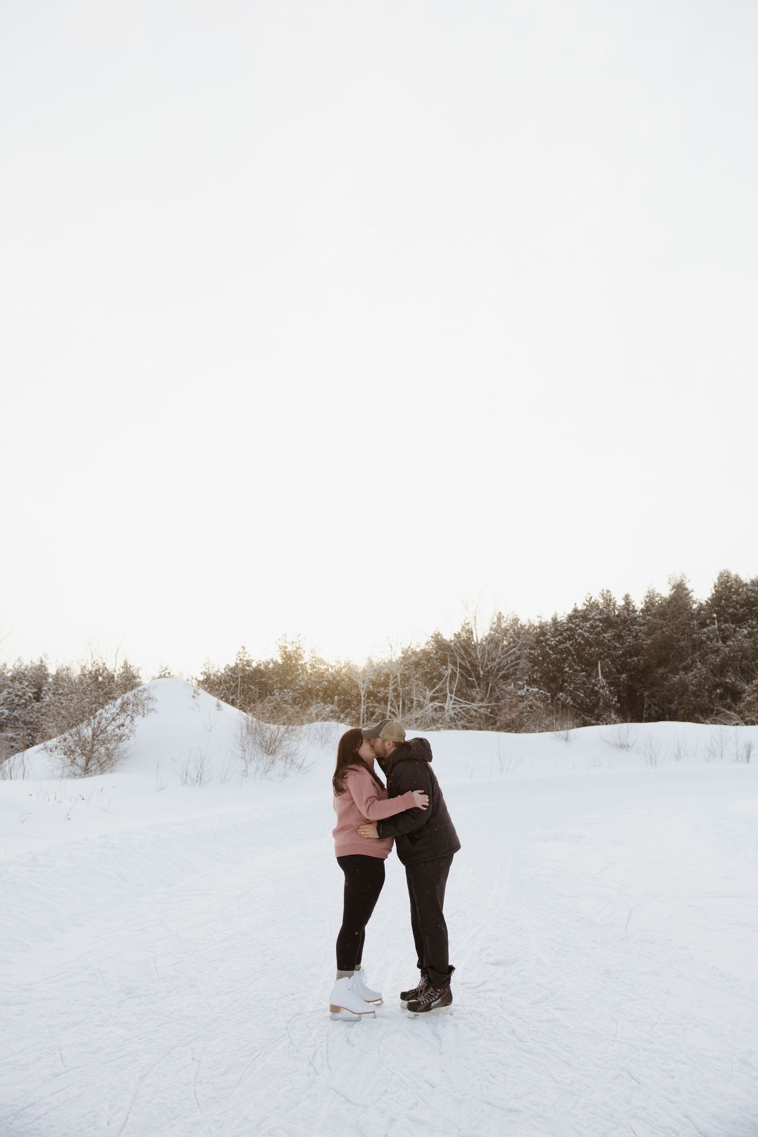 A couple shares a romantic kiss on a frozen lake surrounded by snow-covered trees at sunset.