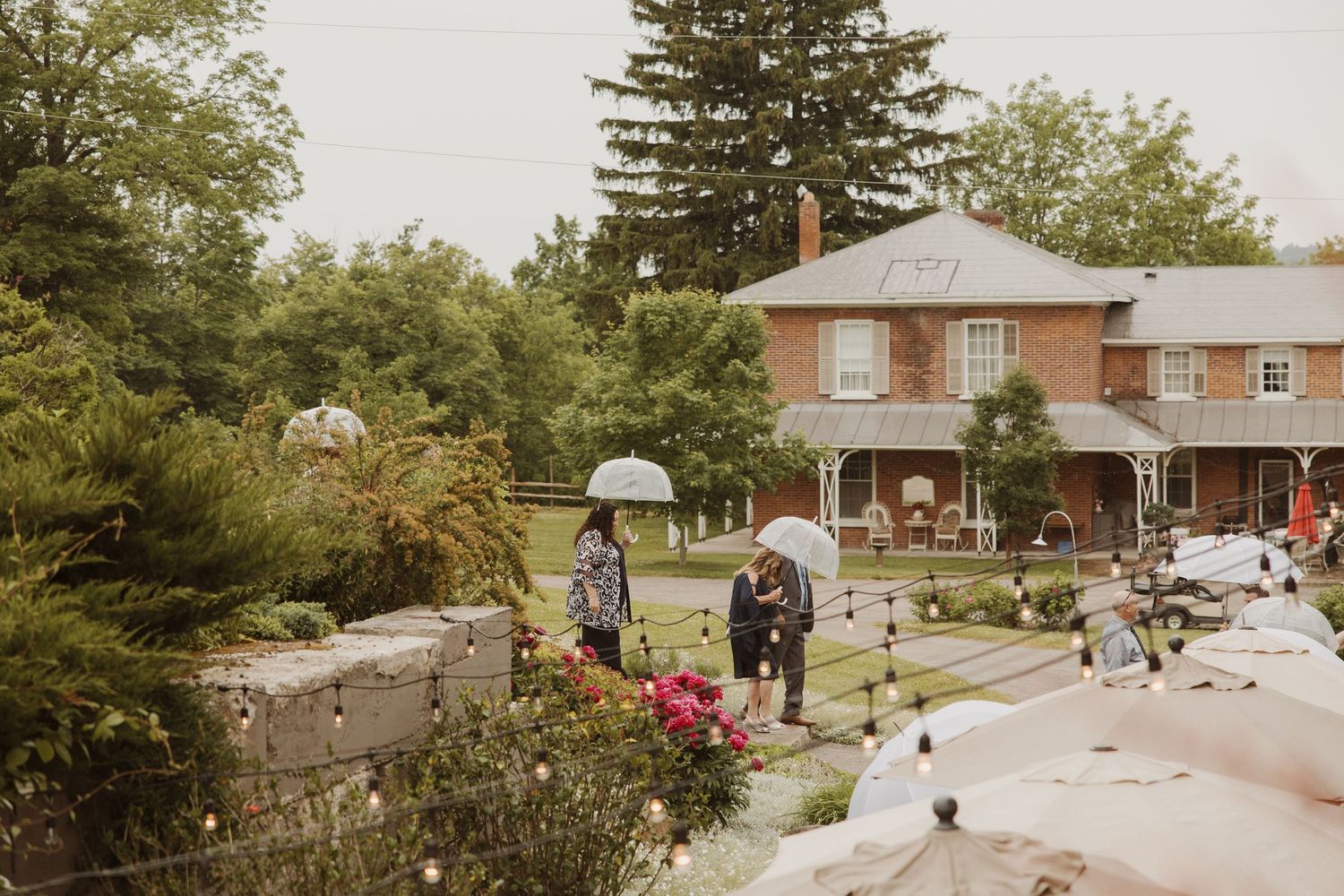 Guests with umbrellas arrive at charming brick farmhouse wedding venue on a rainy day.