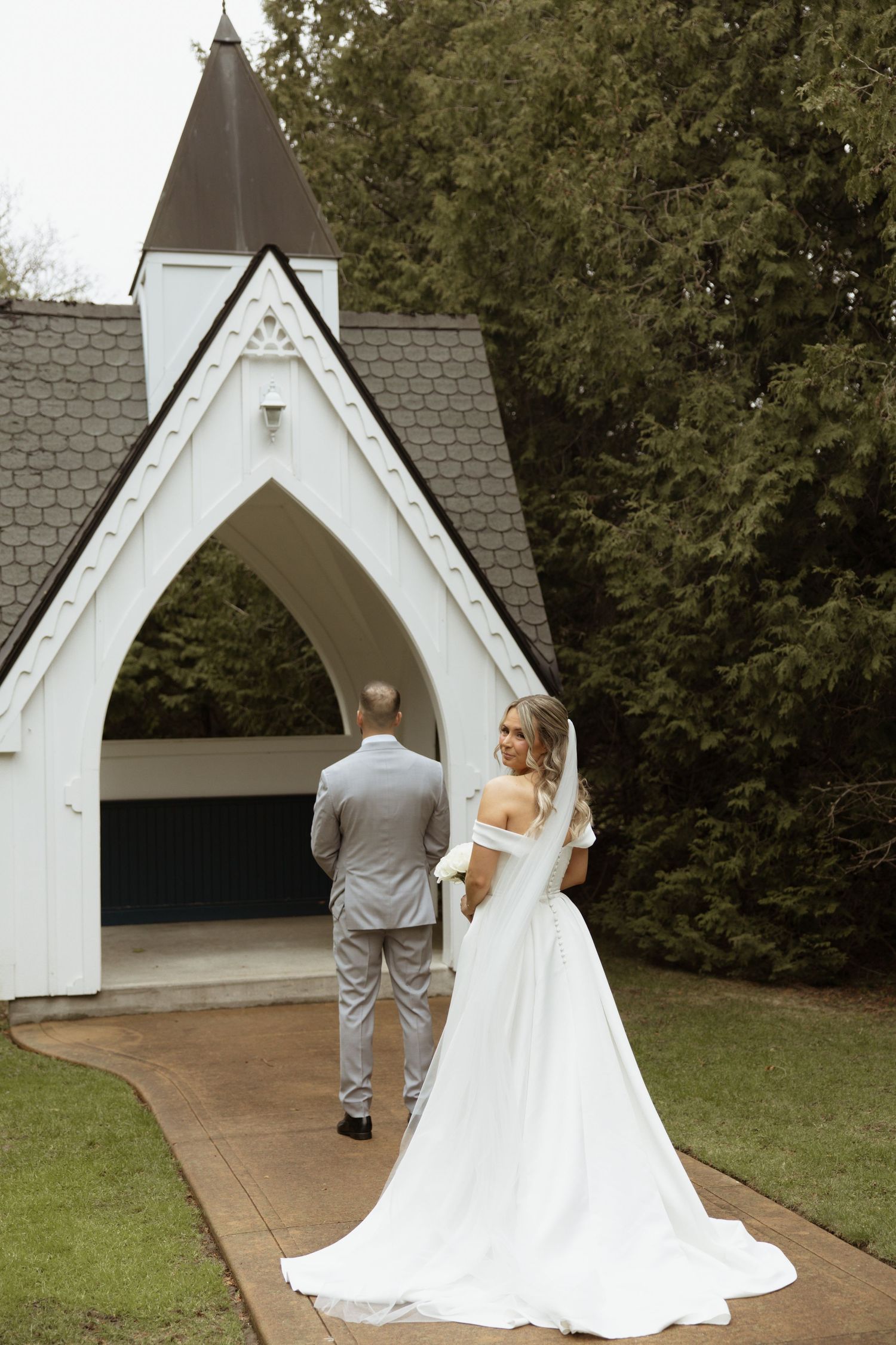 Bride in white gown with train and groom in gray suit stand before white chapel arch surrounded by lush greenery.