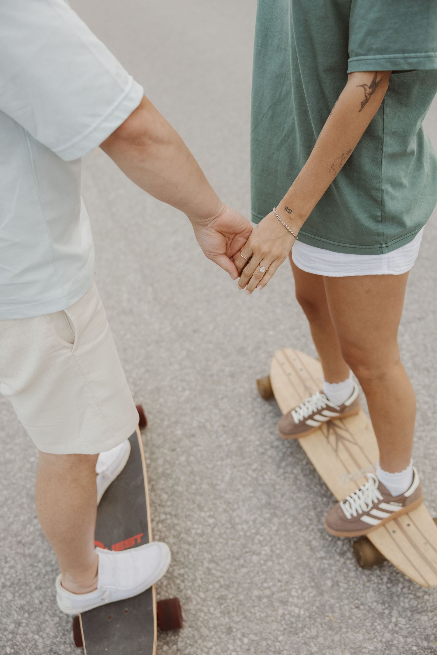 Couple holding hands while riding skateboards on pavement, engagement ring visible.