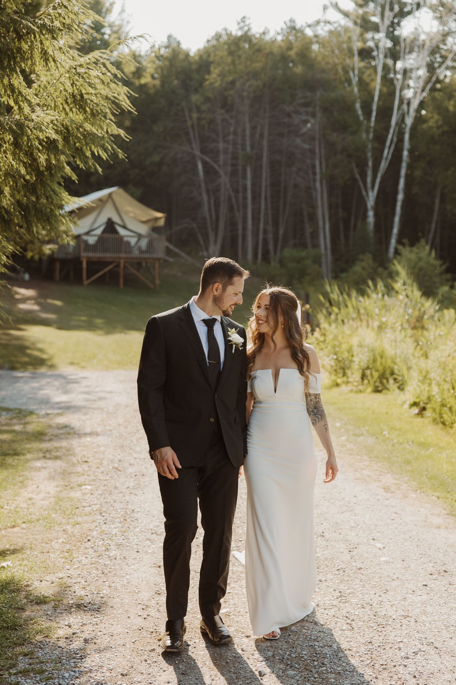 Bride and groom walking together on a sunlit gravel path surrounded by lush green forest.
