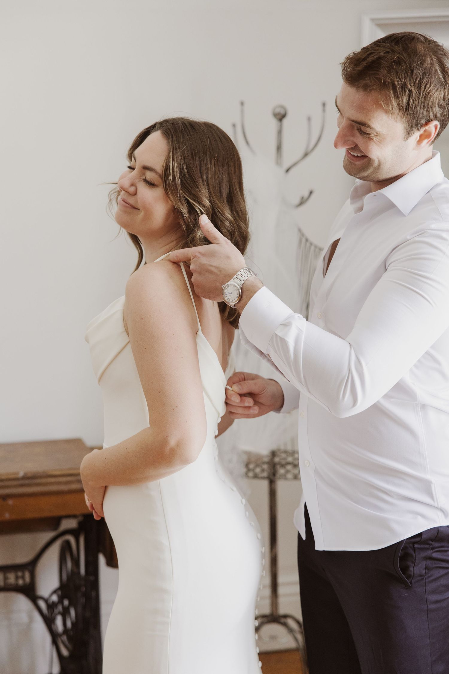 Smiling groom fastens necklace on bride in white slip dress during wedding preparations.