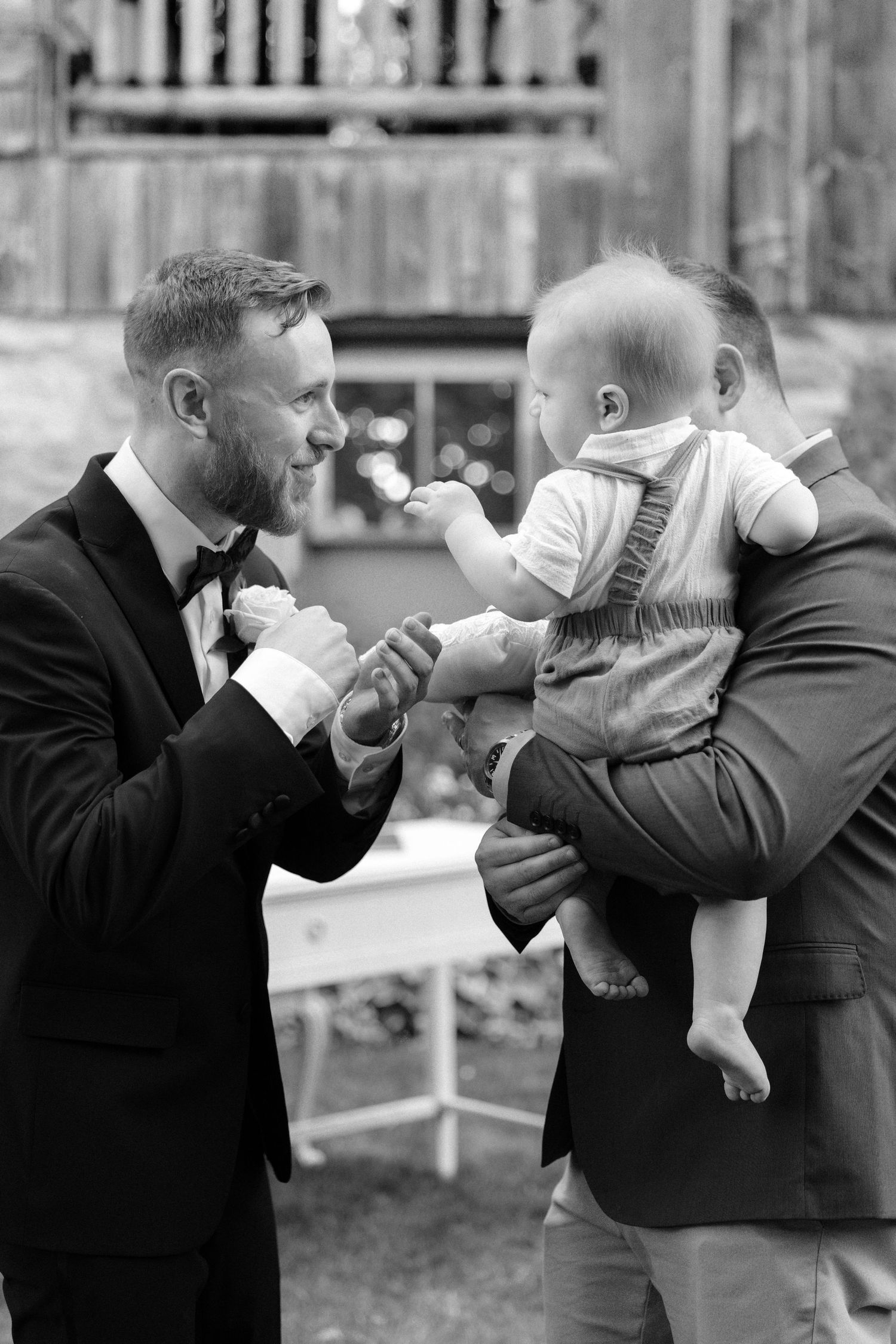 Black and white photo of a groom in a tuxedo interacting with a baby at a wedding.