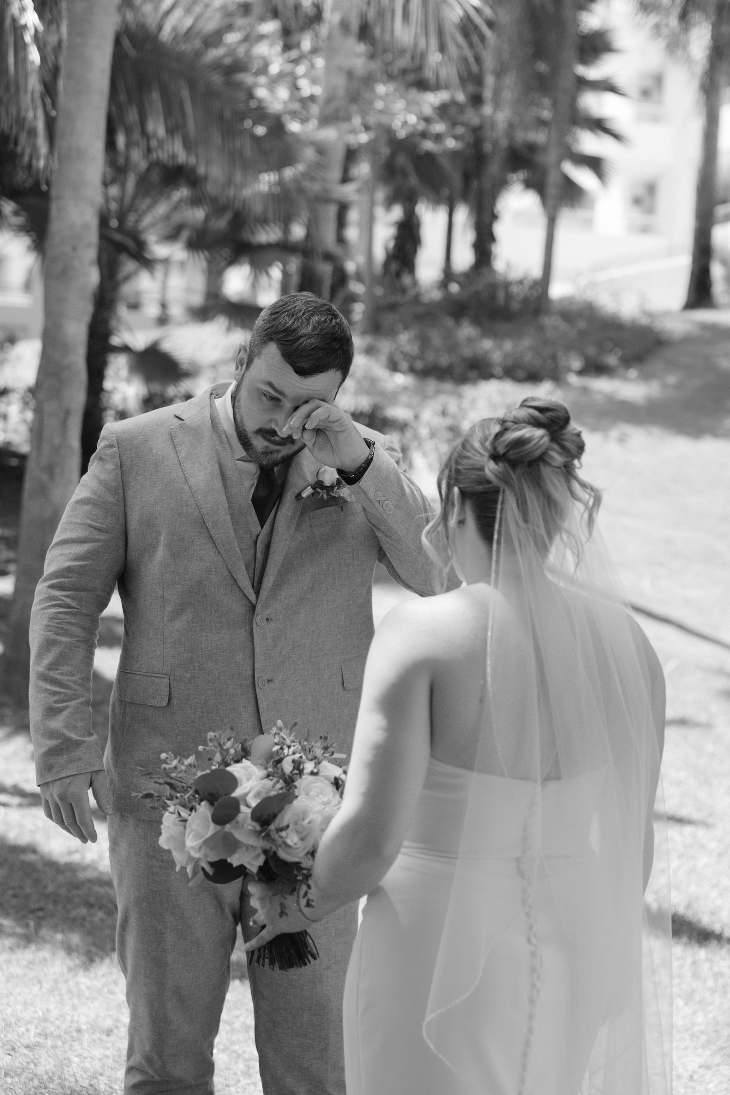 Emotional groom wipes tears as bride approaches during first look moment outdoors in black and white.