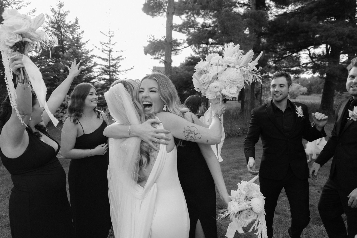 Black and white photo of joyful bride embracing wedding party guests outdoors with bouquets raised.