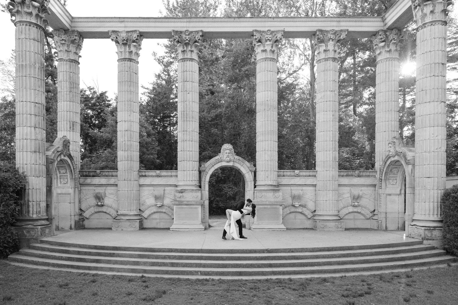 Black and white photo of couple embracing under ornate stone colonnade with arched entrance and decorative columns.
