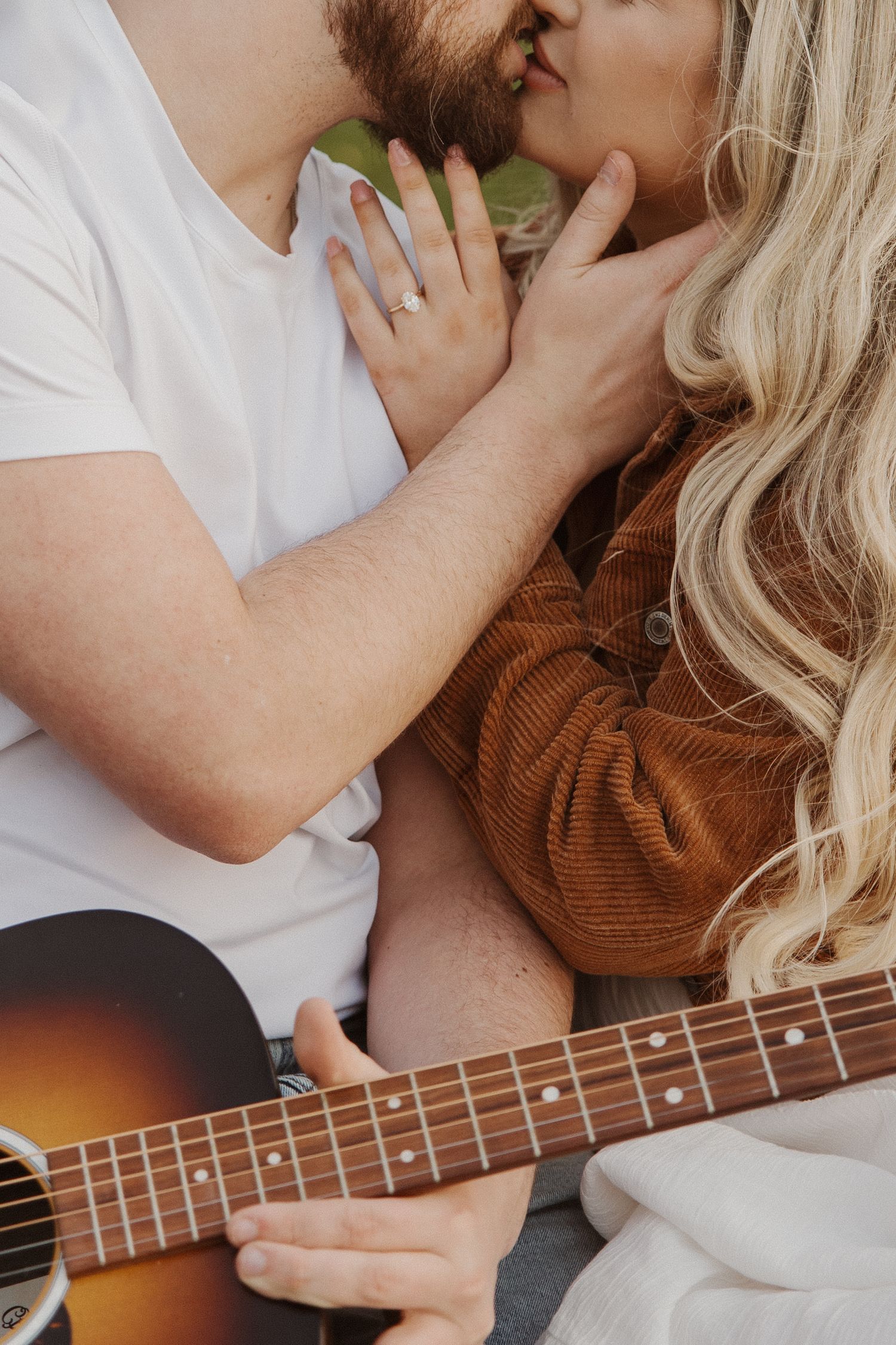 Engaged couple kissing while man holds acoustic guitar outdoors.