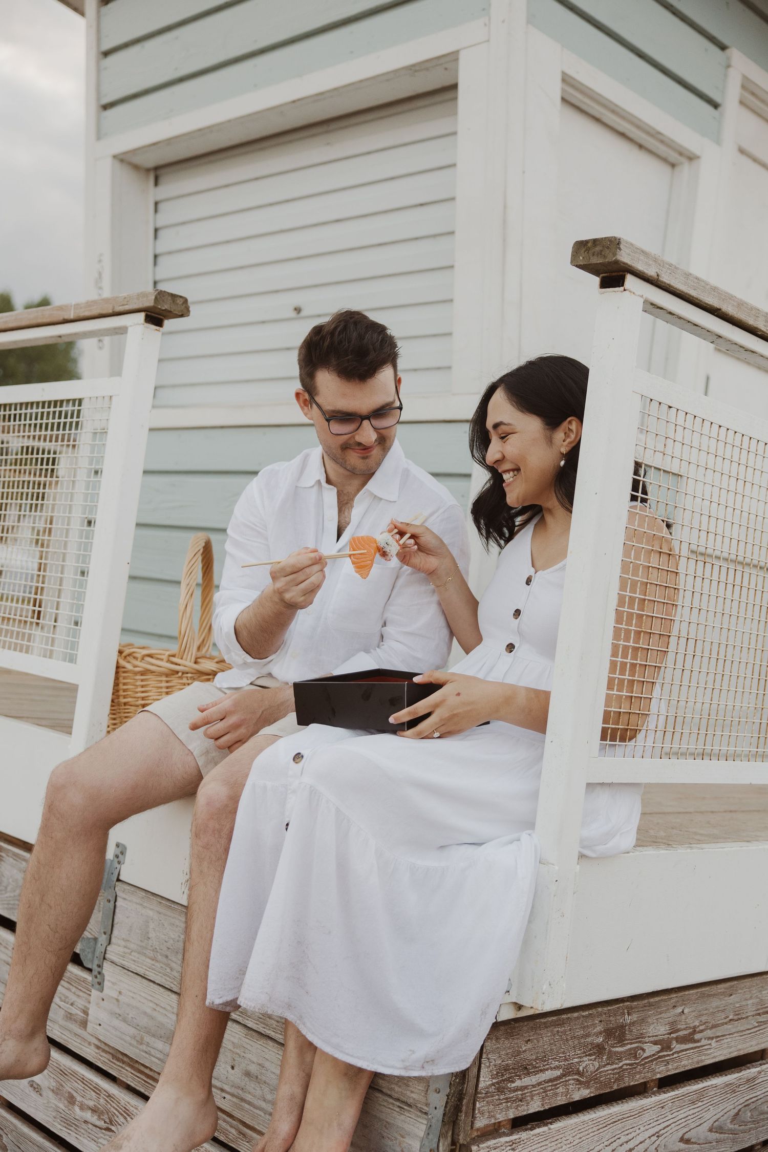 Couple in white outfits sharing a snack on wooden beach hut steps with a wicker basket nearby.
