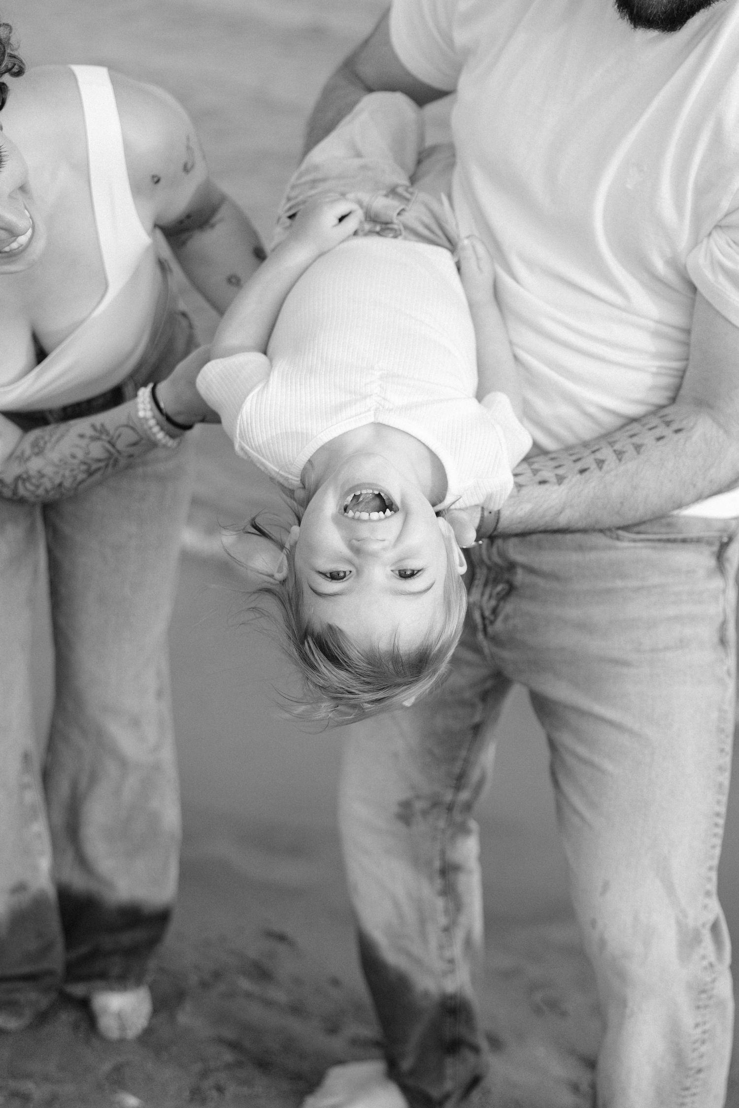 Black and white photo of parents playfully holding their laughing baby upside down at the beach.