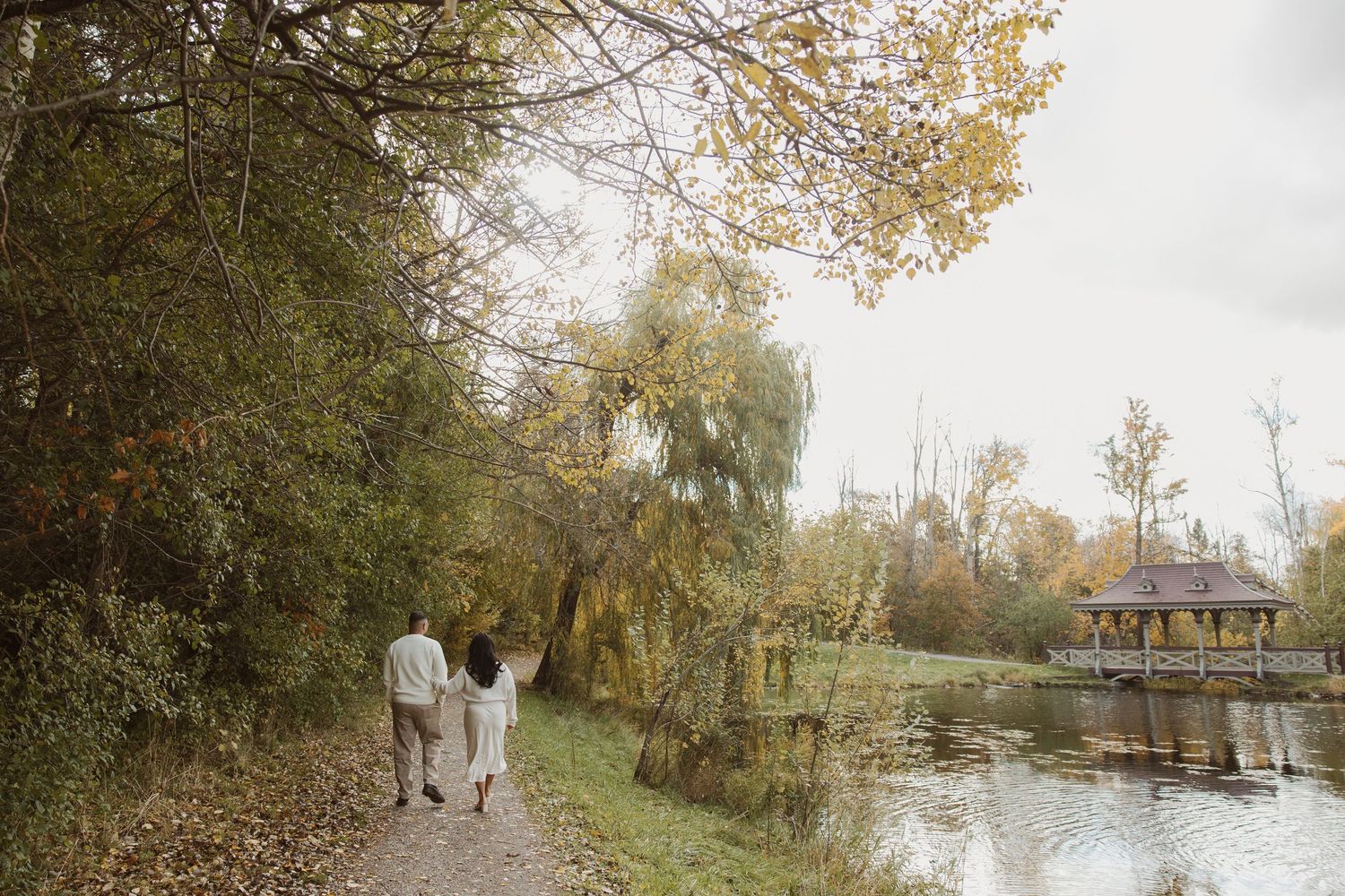 Couple walking hand in hand along a scenic autumn path beside a calm pond with a gazebo in the background.