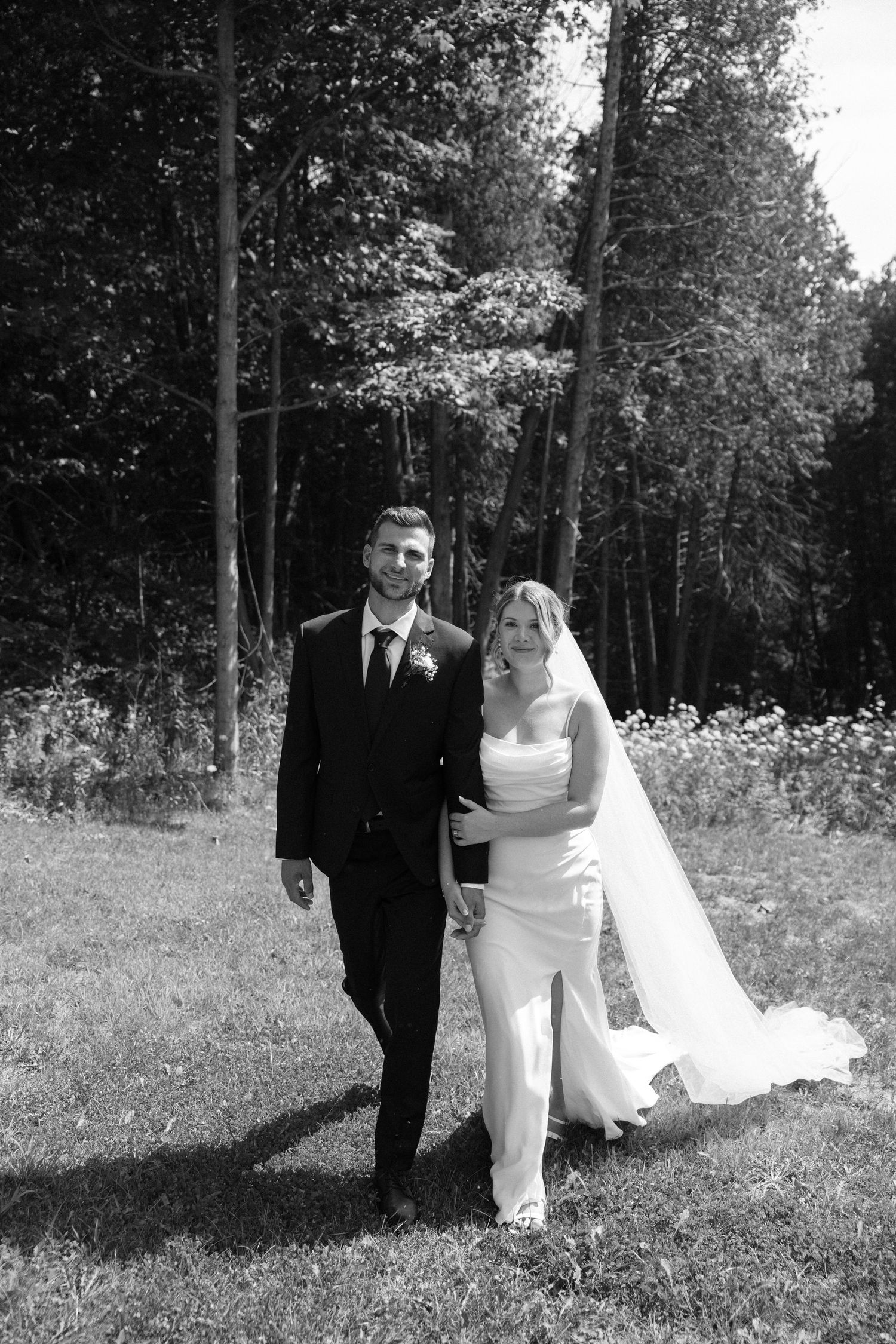 Black and white wedding portrait of bride and groom walking together on sunny grassy lawn.