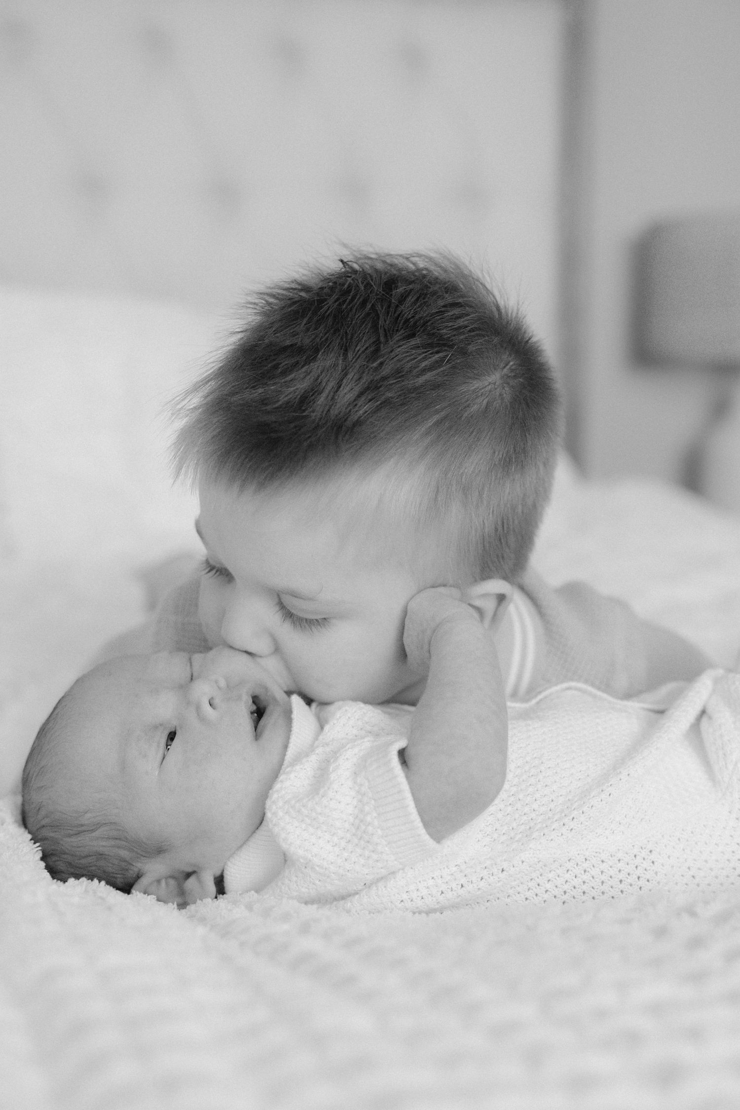 Black and white photo of a young boy gently kissing his newborn sibling on a bed.