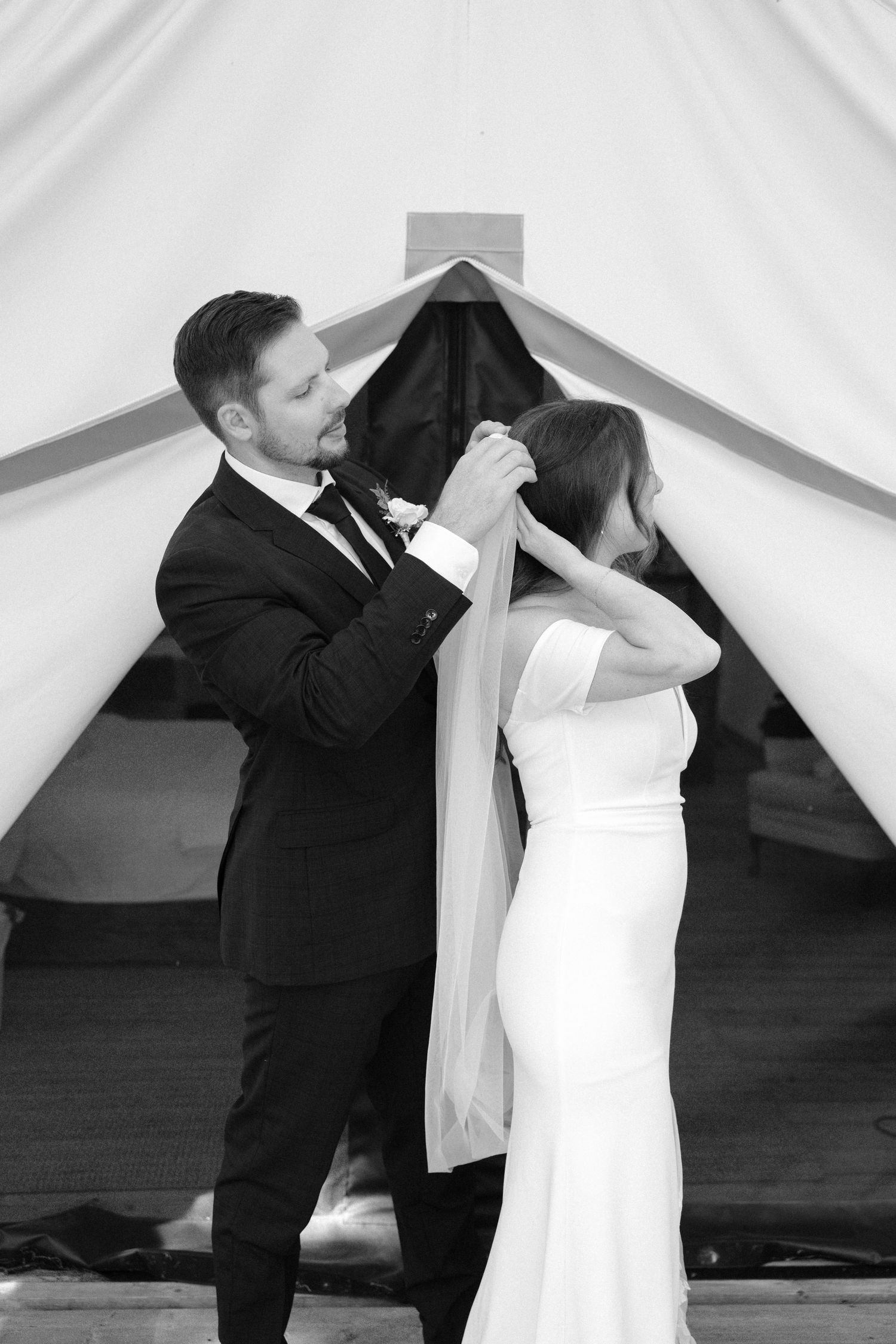 Groom placing veil on bride in black and white wedding photo under a draped tent.