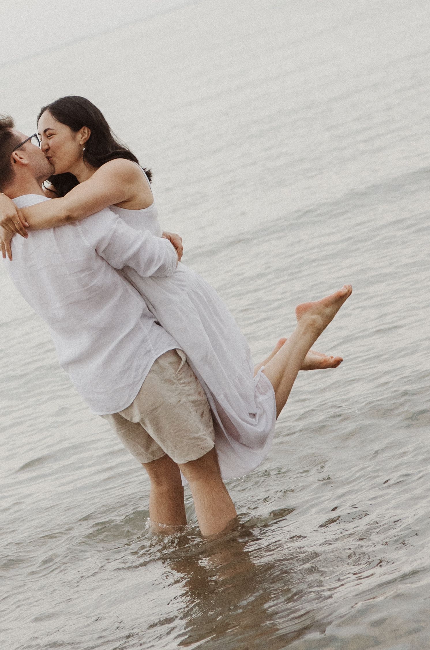 Romantic couple kissing as man lifts woman in his arms while standing in shallow ocean water.