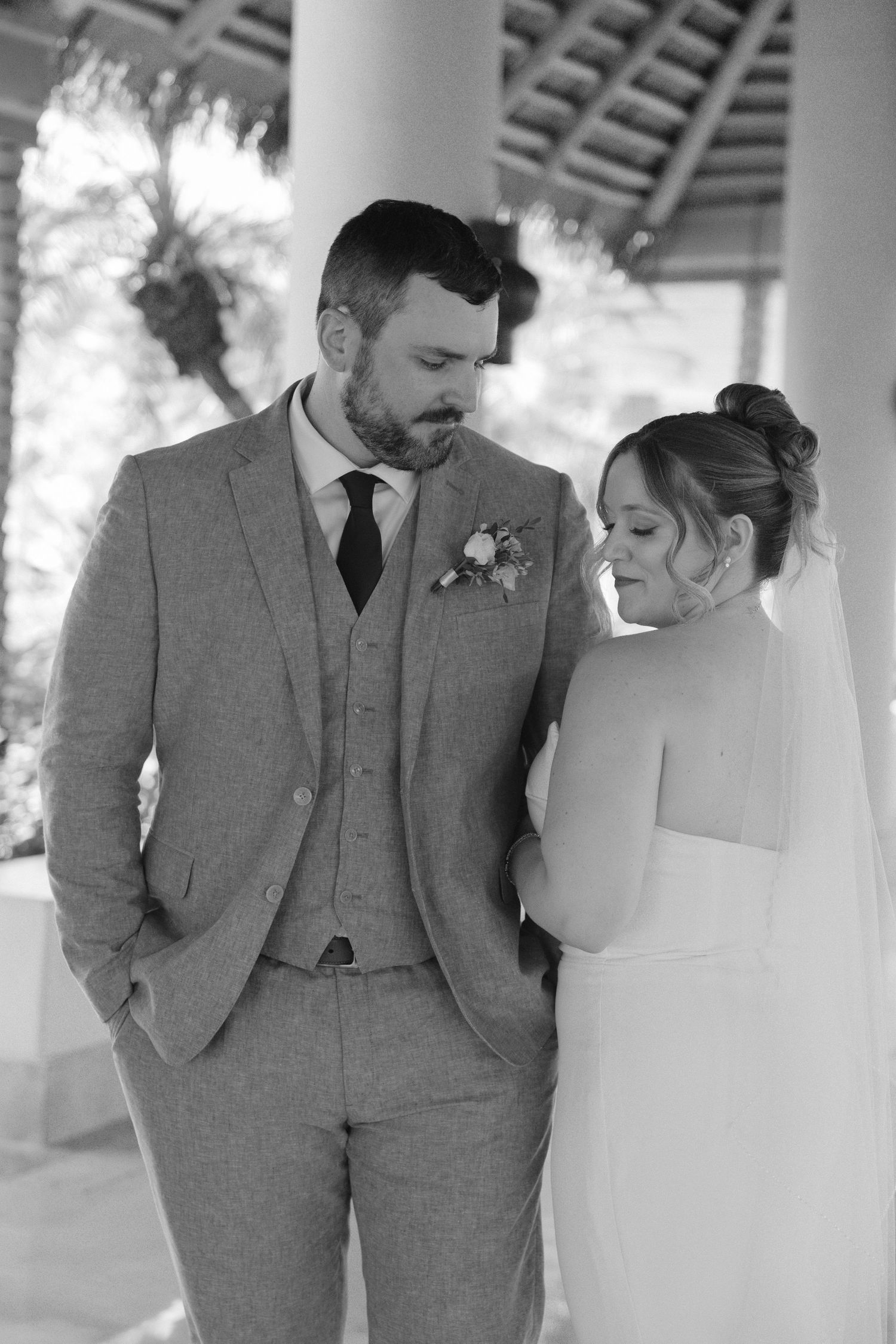Black and white photo of bride and groom in elegant wedding attire sharing a tender moment under a covered outdoor venue.