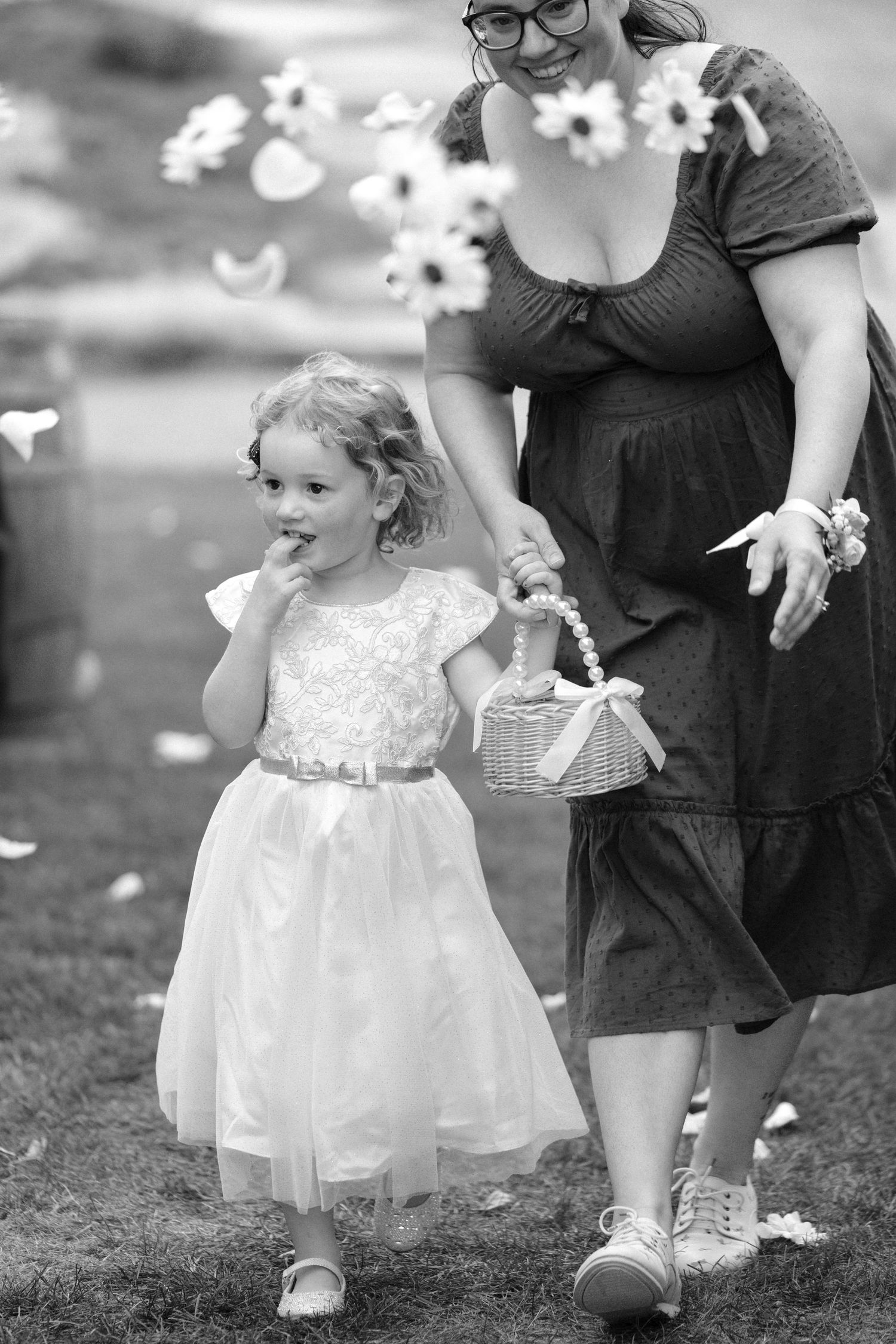 Black and white photo of a flower girl in a white dress walking with a woman at a wedding.