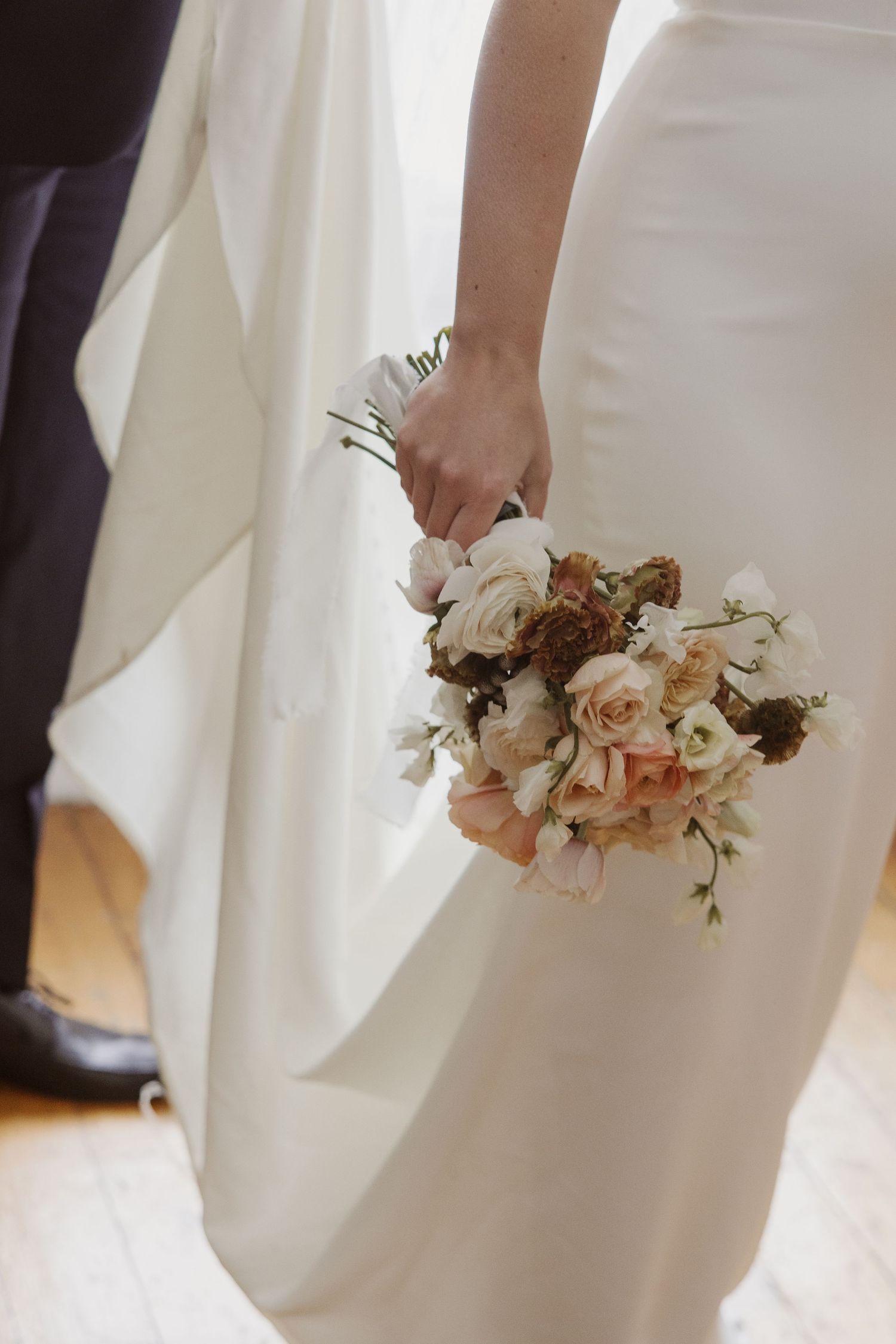 Bride in sleek white gown holds delicate bouquet of blush and ivory roses at wedding.
